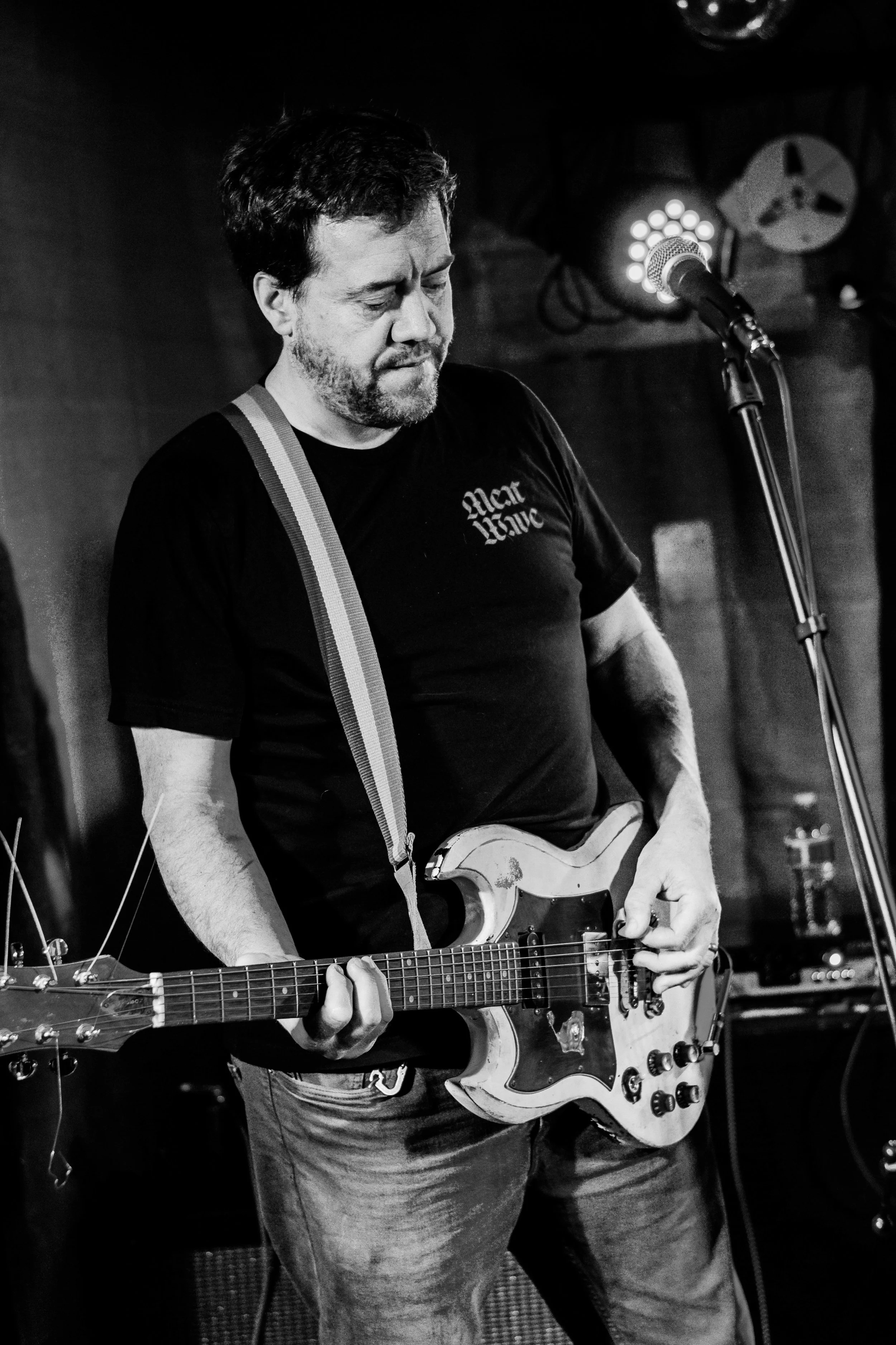 A man playing an electric guitar on stage, focused on his performance, with stage lights and equipment in the background, in black and white.