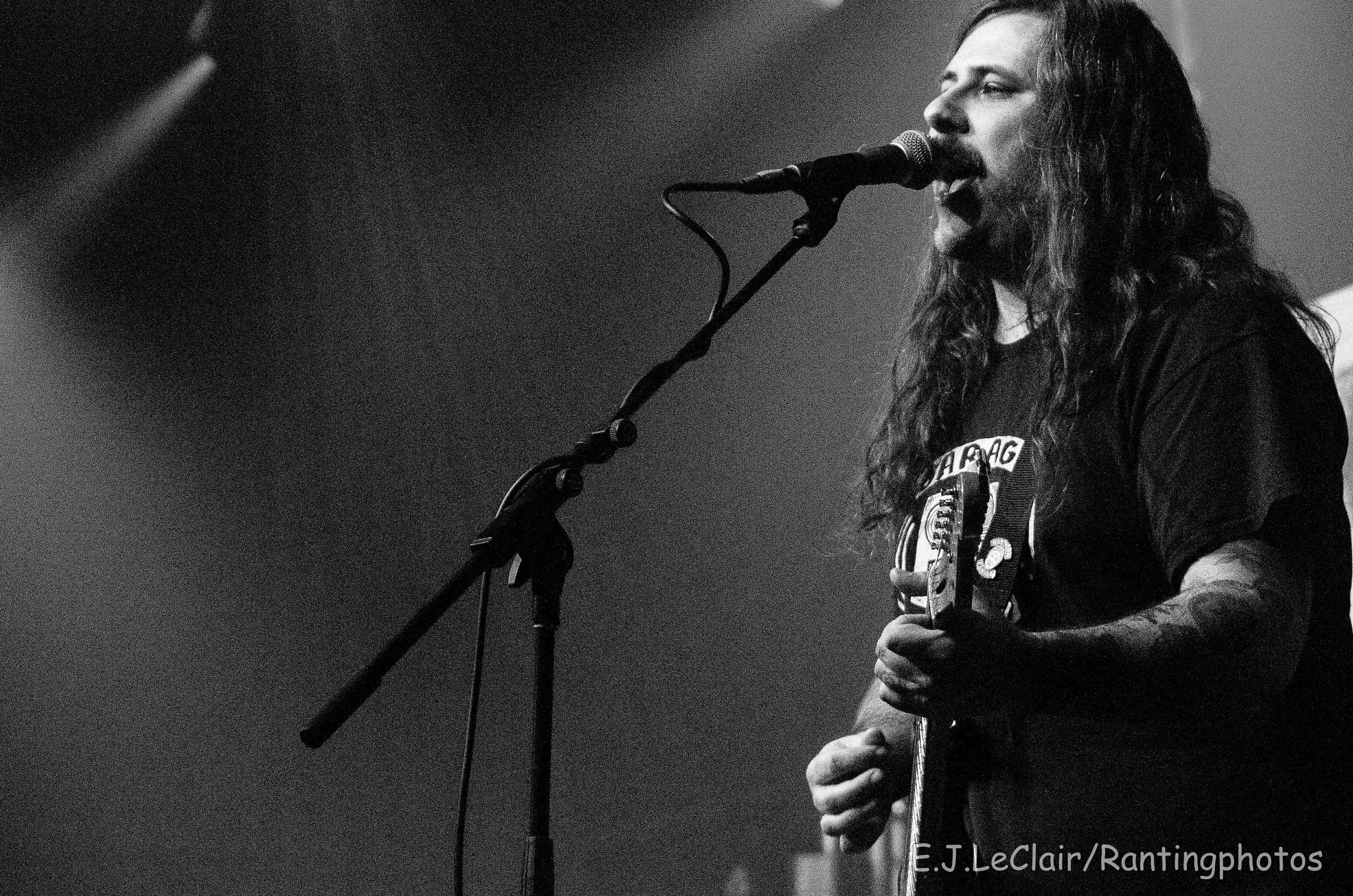 A person with long hair and a mustache singing into a microphone while holding a guitar on a stage.