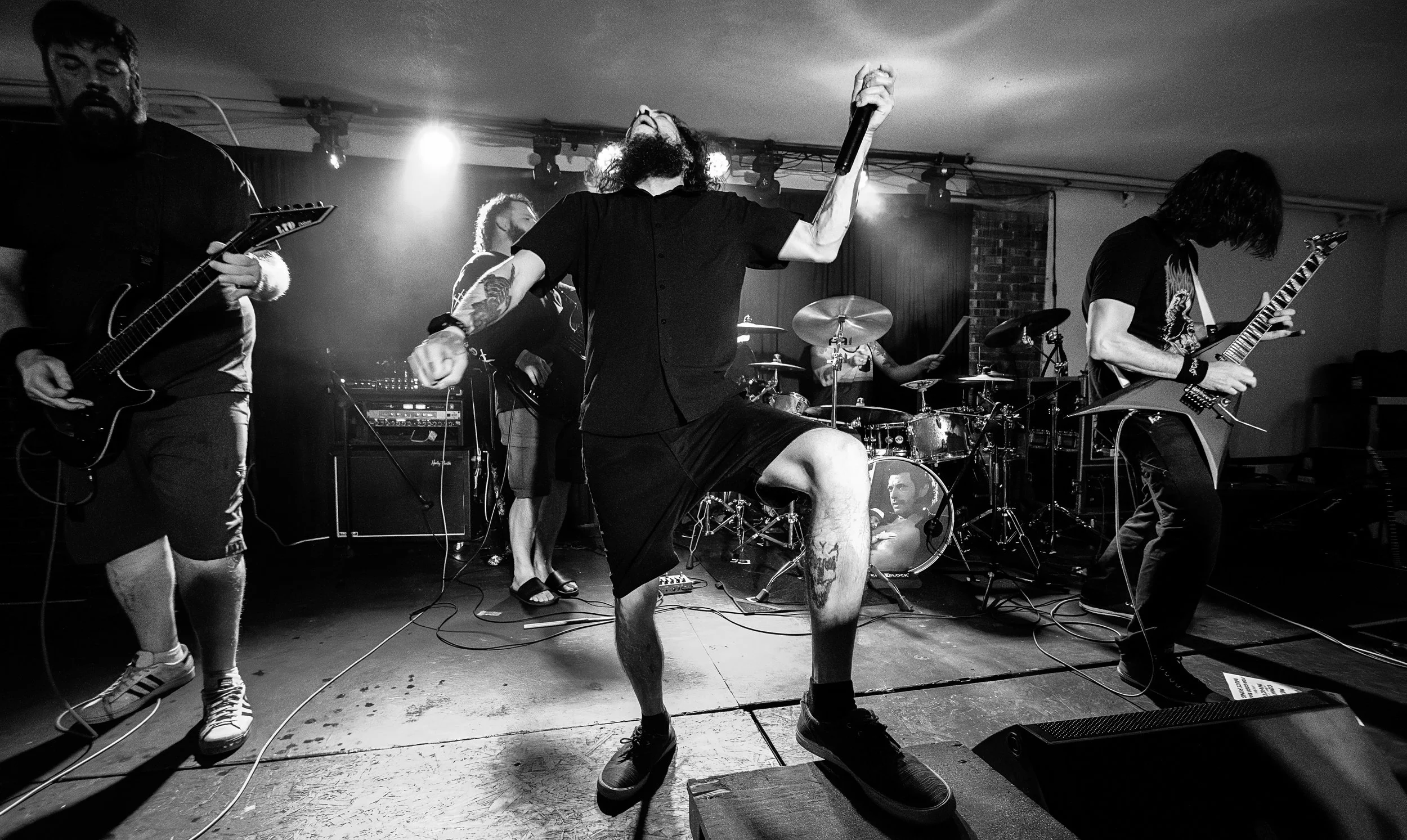 Black and white photo of a band performing on stage, with a vocalist in the center holding a microphone, accompanied by guitarists and drummer.