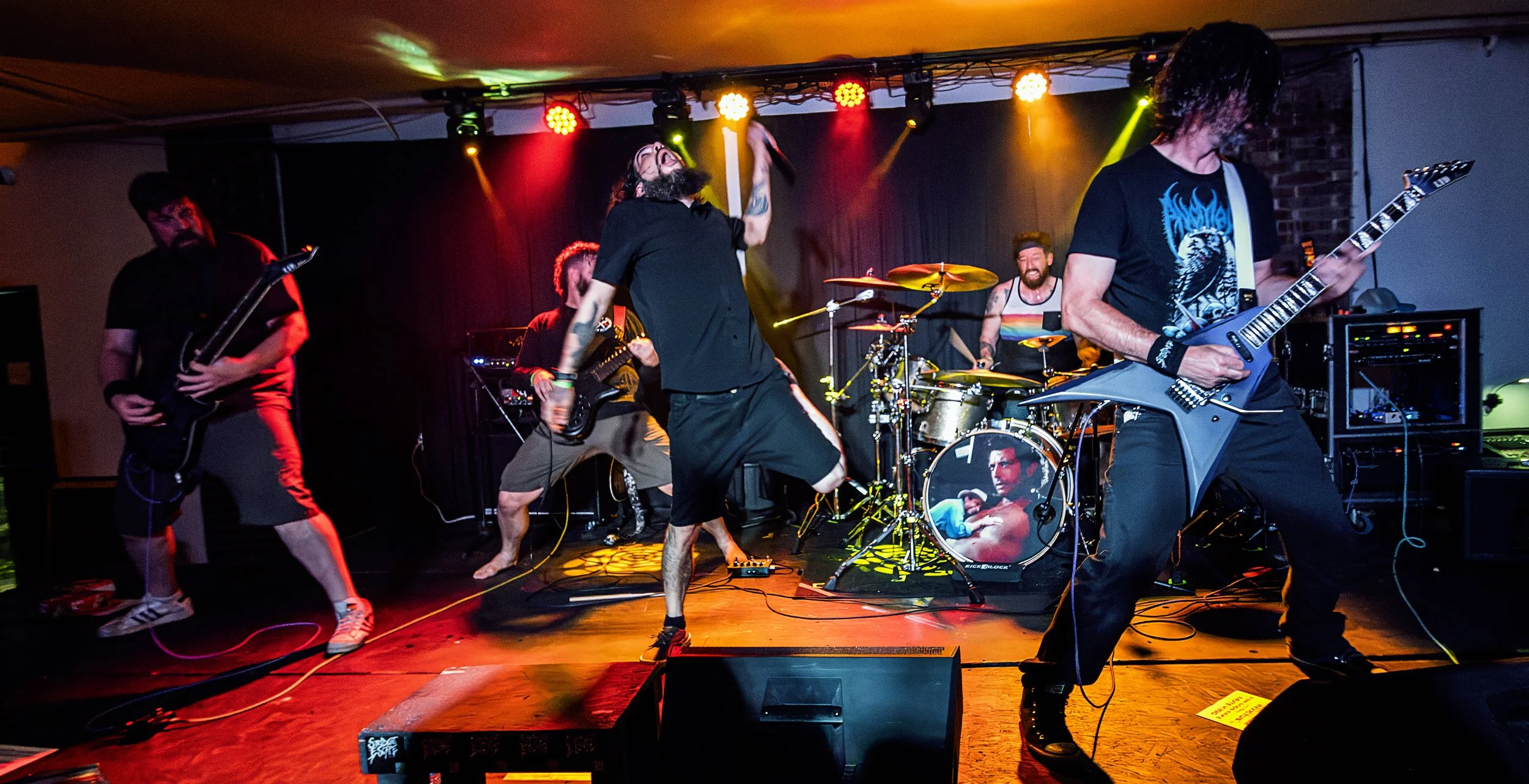 A band performing live on stage with multiple musicians playing guitars, drums, and singing, illuminated by colorful stage lights.