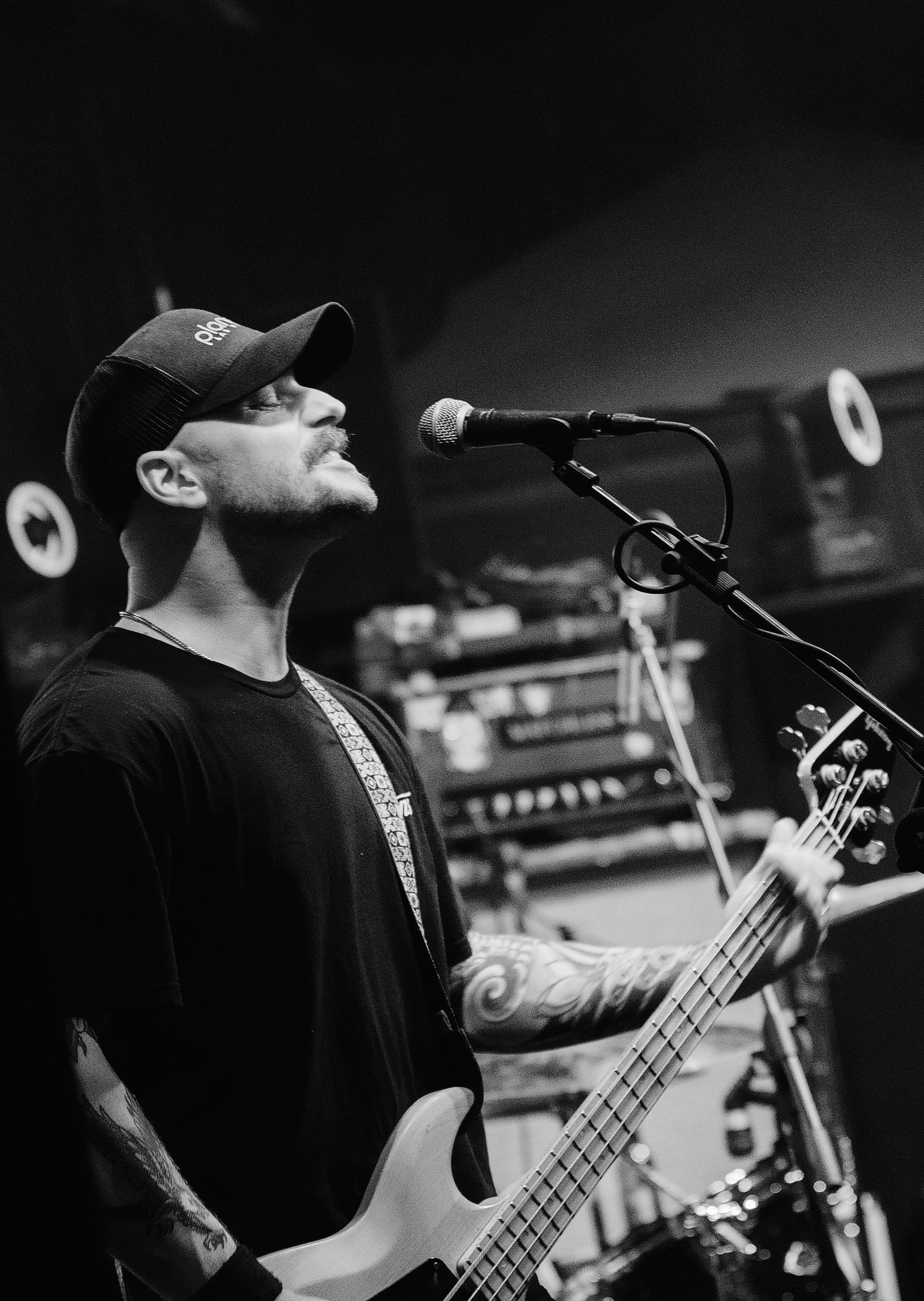 A man wearing a baseball cap and black T-shirt, singing into a microphone while playing an electric bass guitar with tattoos on his arm, in a music rehearsal space.