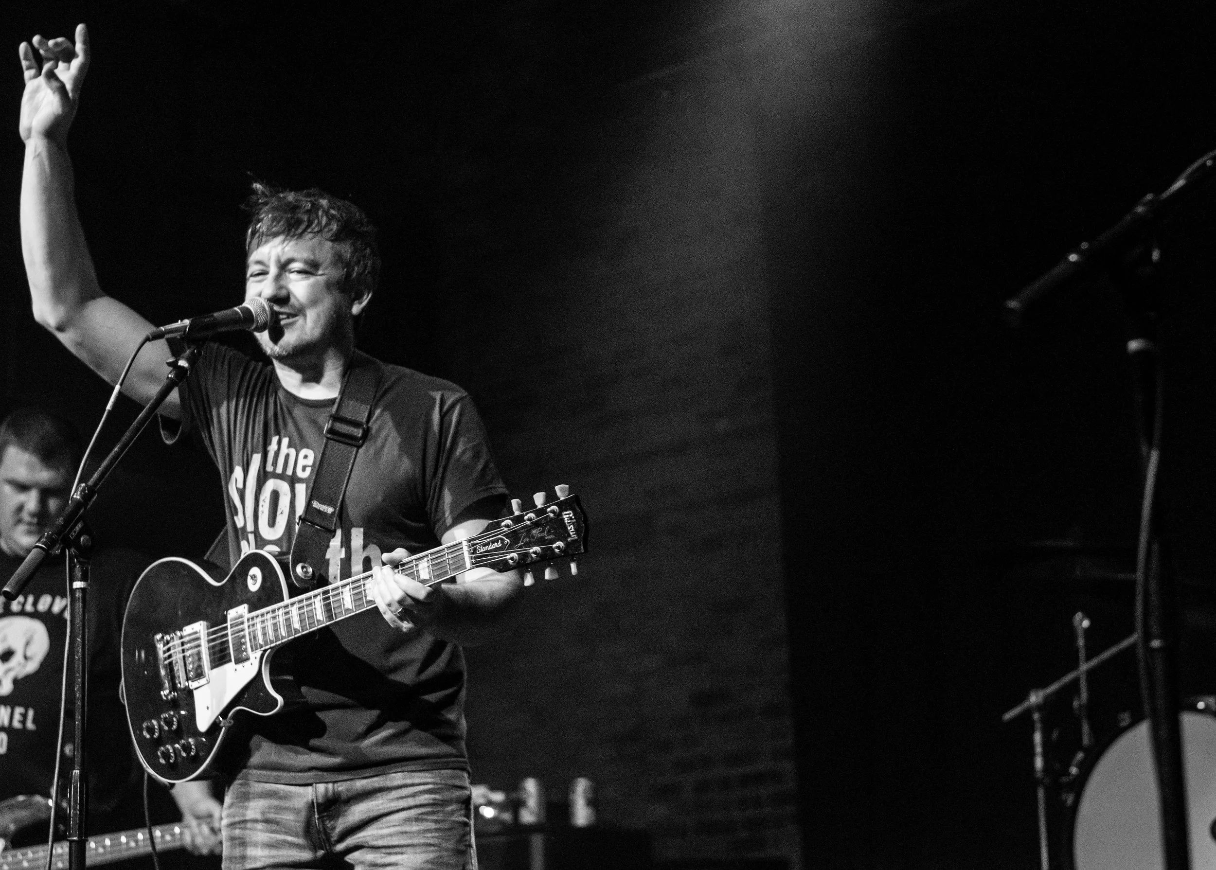 A male musician with short hair singing into a microphone and playing an electric guitar on stage, in black and white.