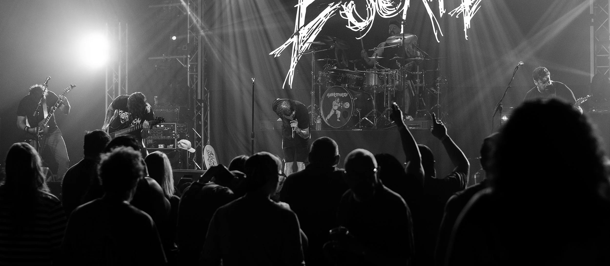 Black and white photo of a live band performing on stage with audience in front, some audience members raising hands.