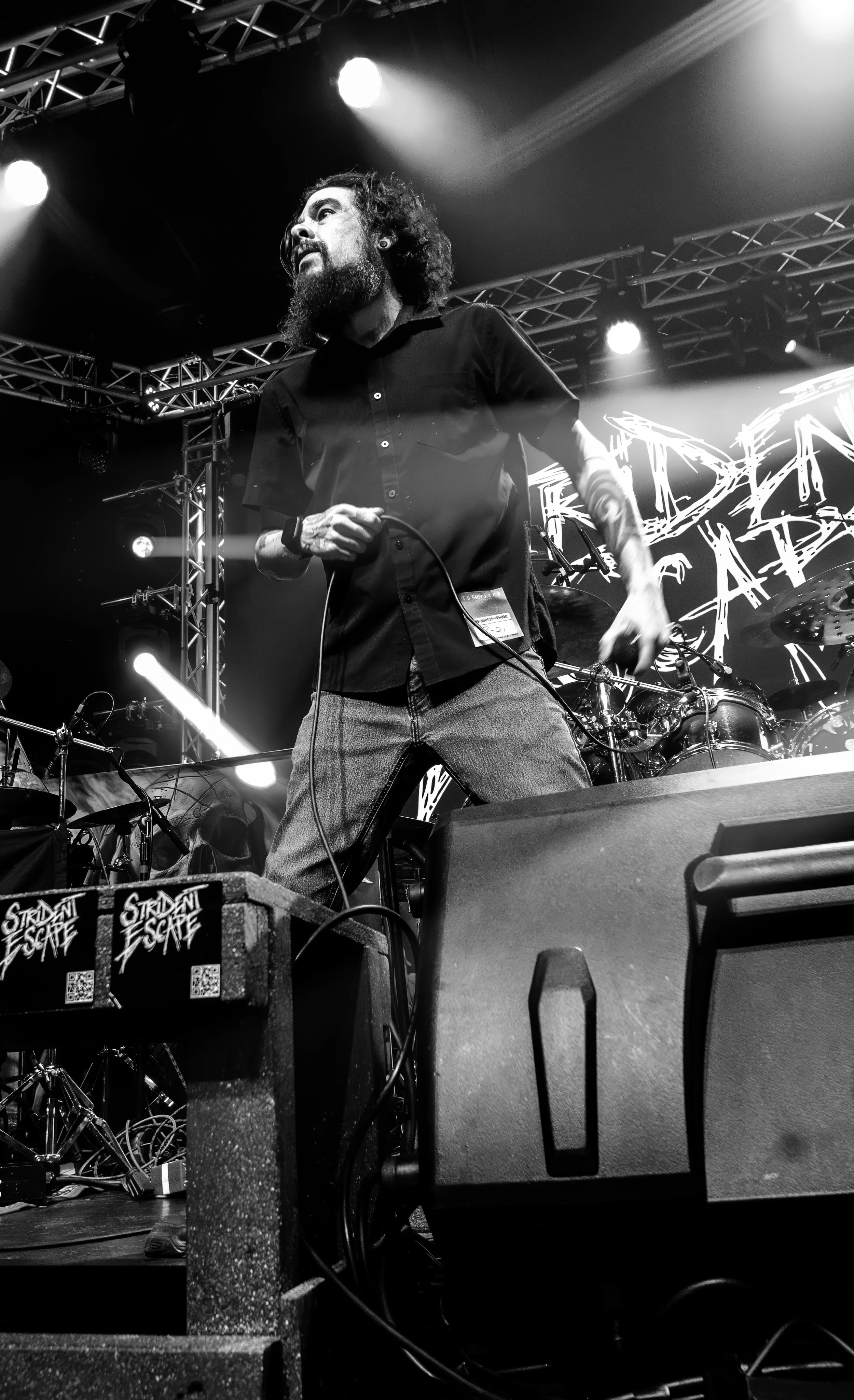 Black and white photo of a musician with a beard and curly hair, wearing a black shirt, performing on stage with stage lights, drums, and equipment, and a backdrop with graffiti-style text.