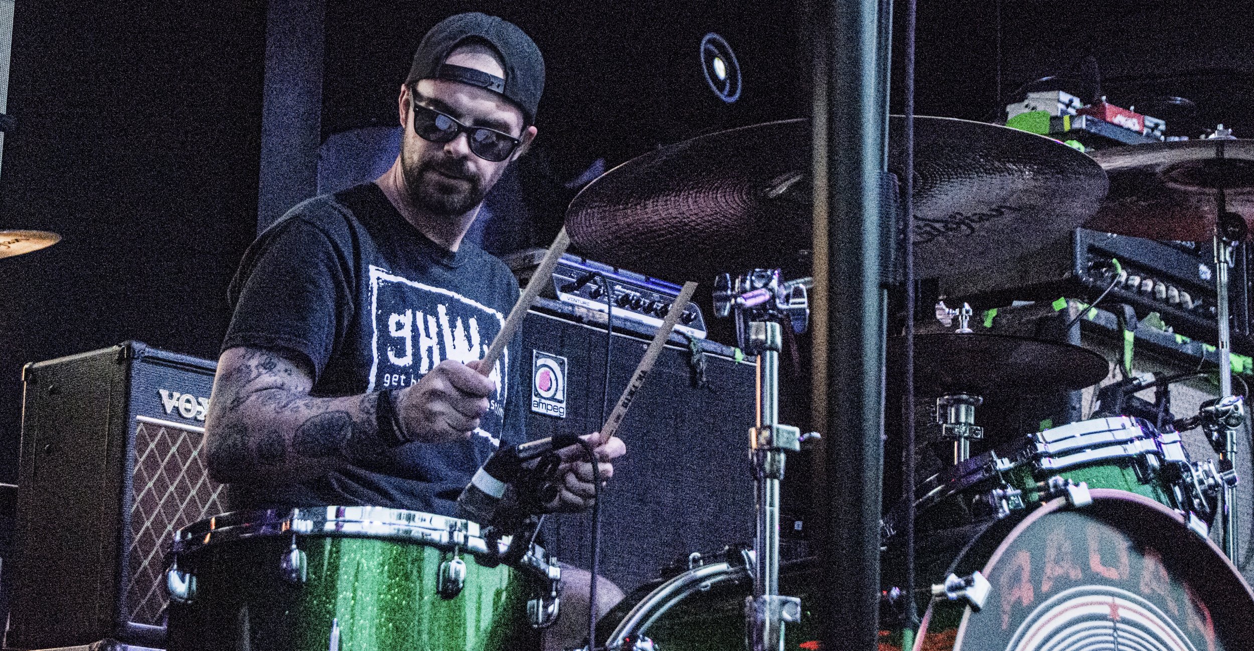 A man with sunglasses and a cap playing drums on stage, surrounded by musical equipment.