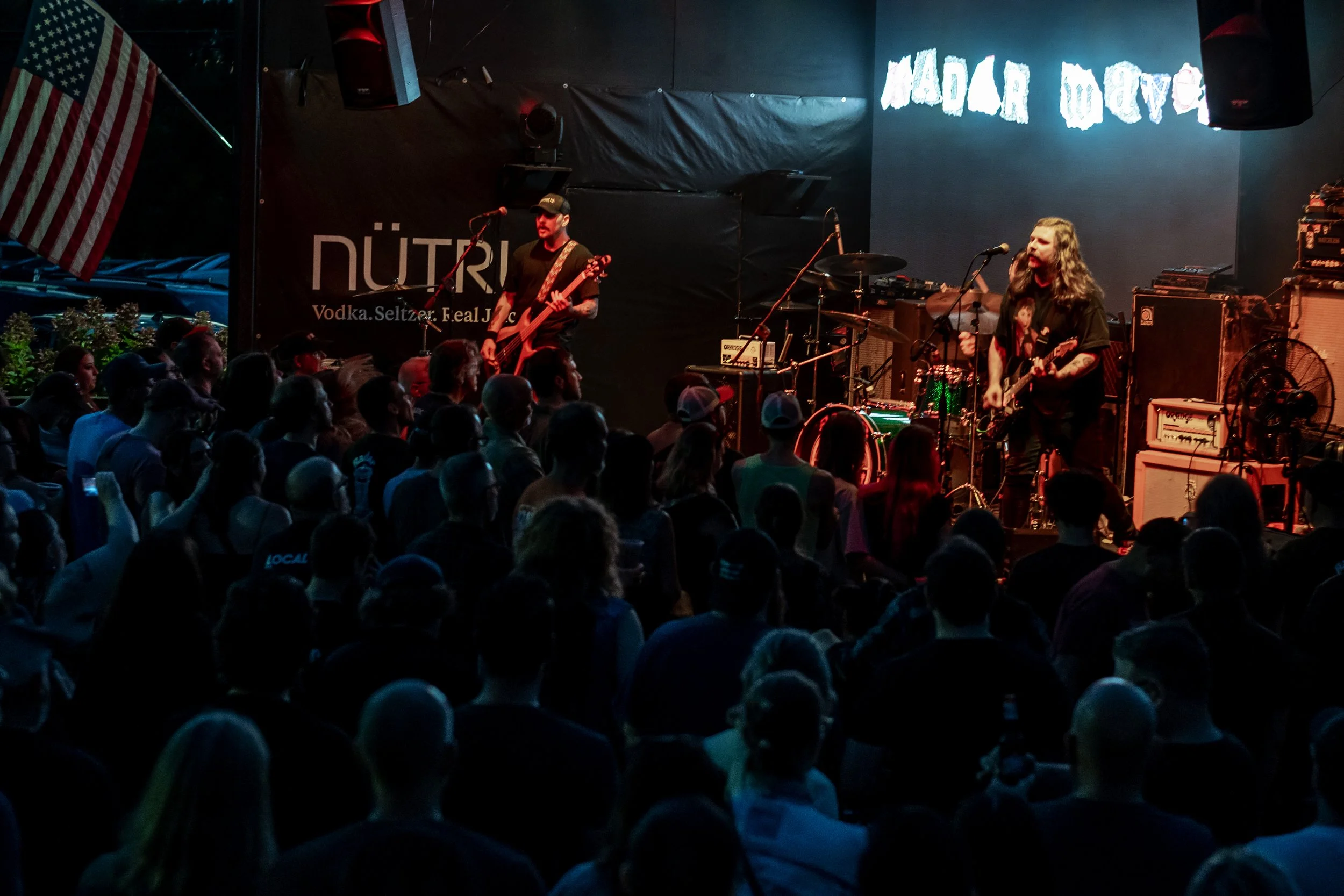 A live music performance on stage with multiple musicians playing guitars and drums, an audience watching, an American flag hanging on the stage, and a lit sign that reads 'MODAR WER'.