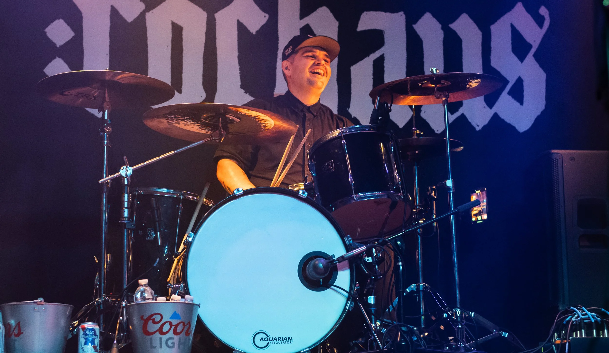 A drummer in a black shirt and cap smiling while playing a drum set on stage. The background has a large graphic with the word 'Faith' and stage lights.