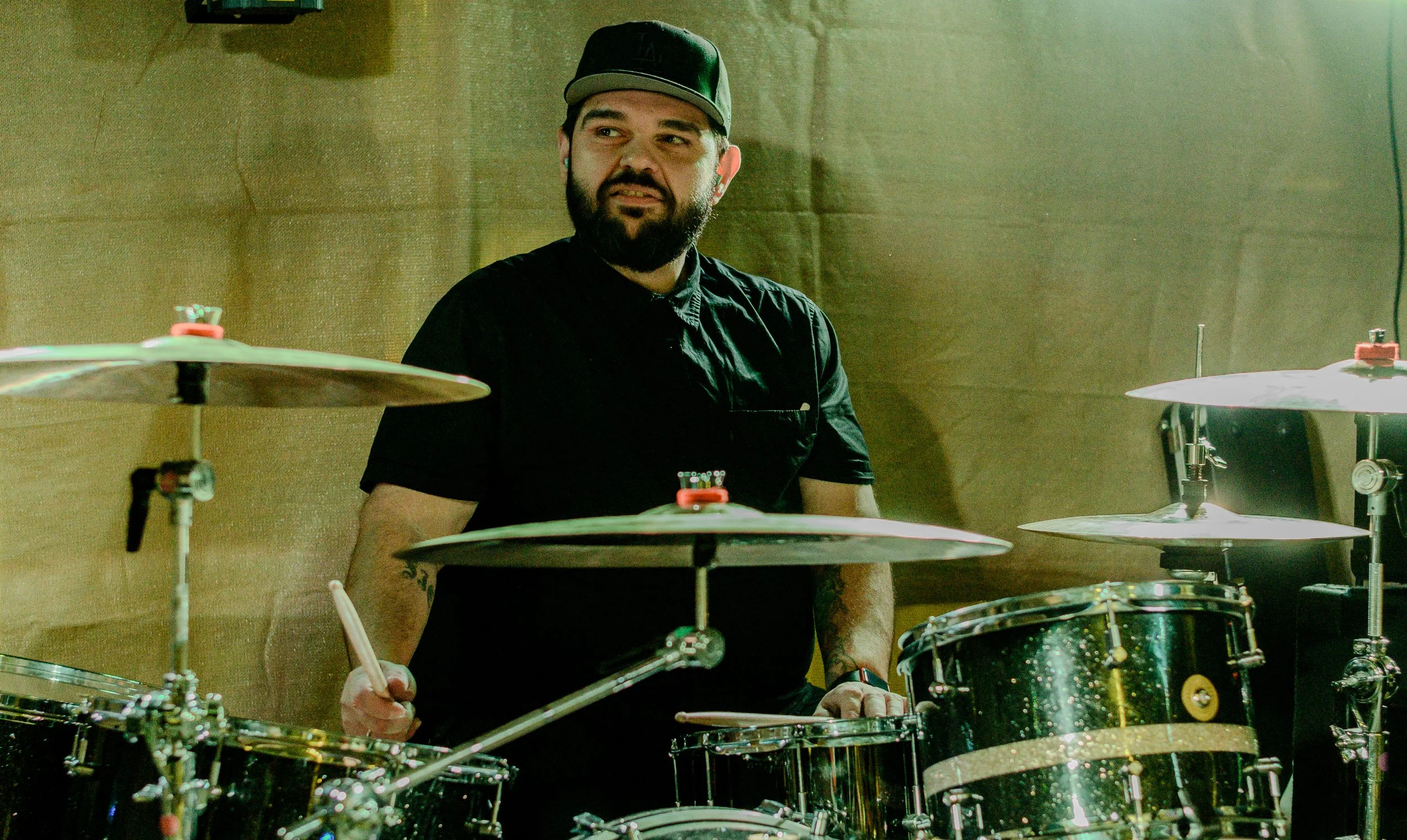 A man with a beard wearing a black cap and black shirt is sitting behind a drum set, holding drumsticks, in a room with a beige wall.