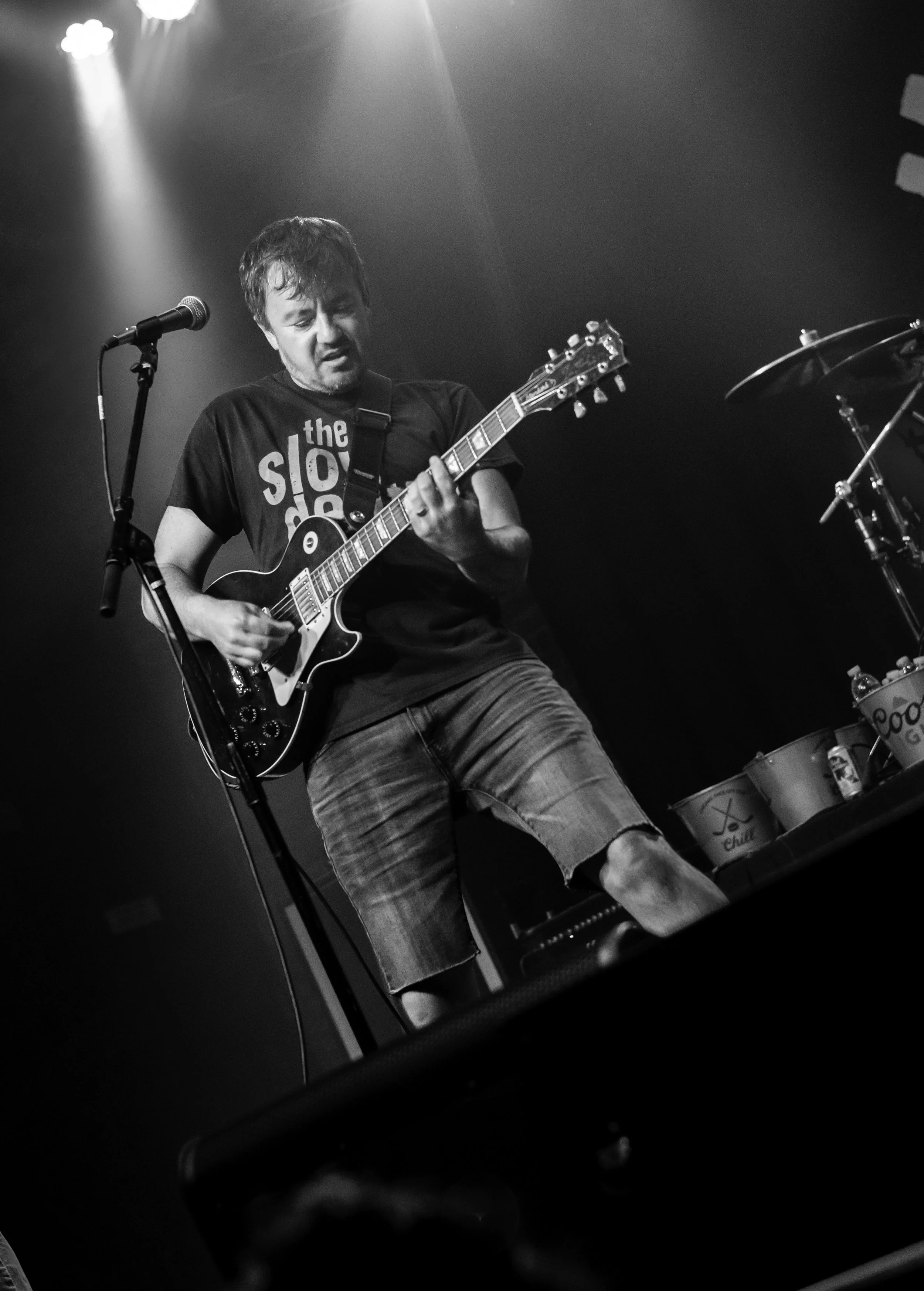 A man plays an electric guitar on stage, wearing a T-shirt with the words 'the slow d' and shorts. He appears to be performing with intensity under stage lights.
