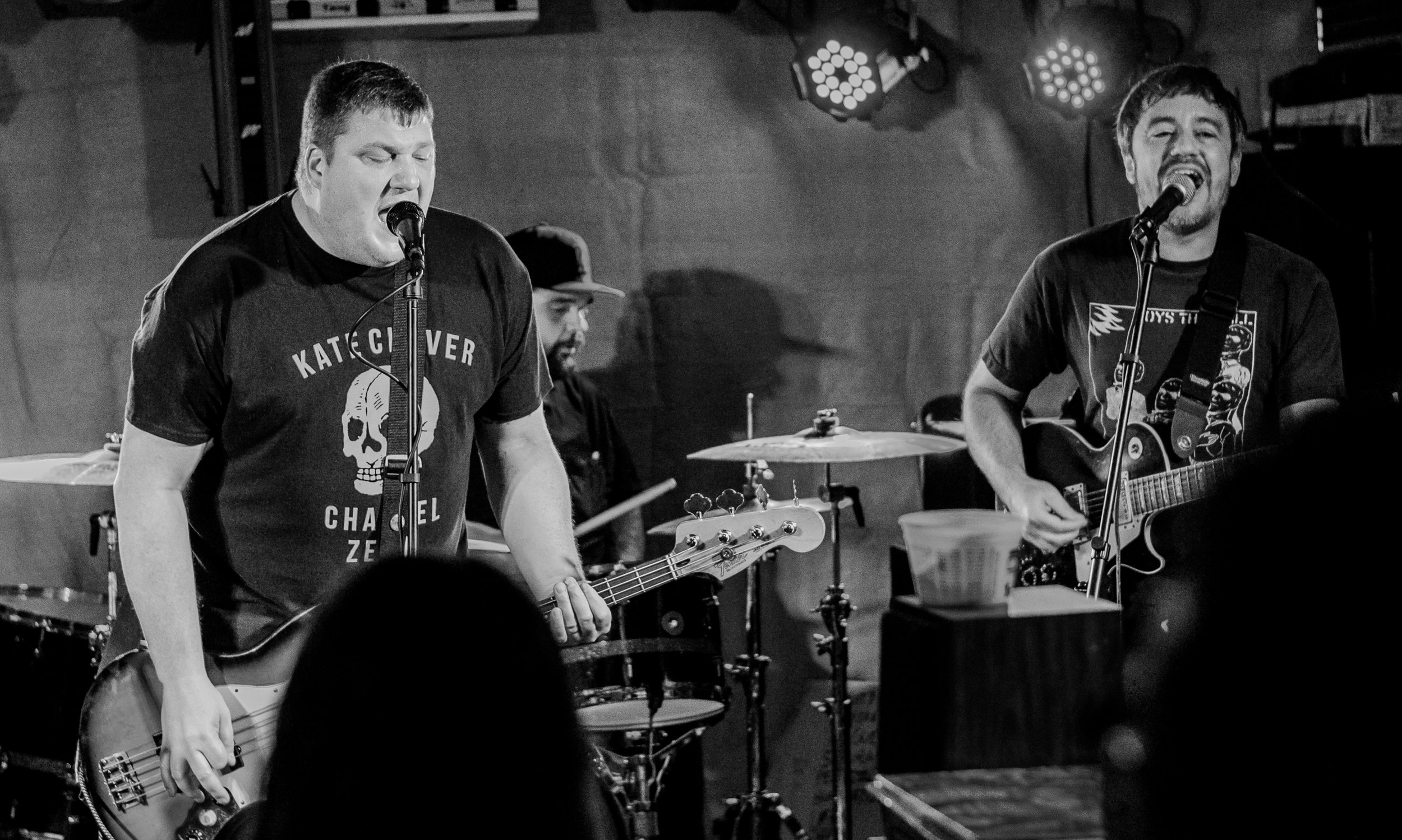 Two male musicians performing on stage, singing into microphones and playing guitars, with a drummer in the background. The scene appears to be at a live music venue, and the image is in black and white.