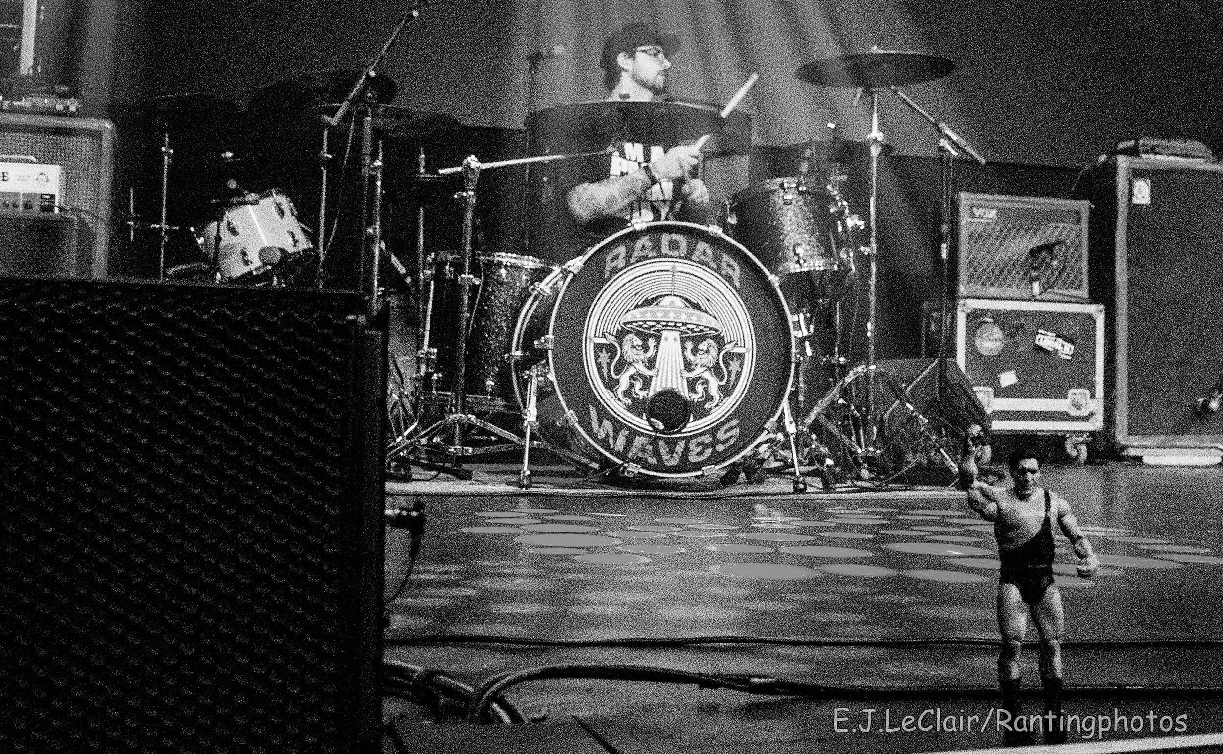 A musician playing drums on stage, with music equipment and speakers around, and a performer speaker standing on the floor in the foreground.