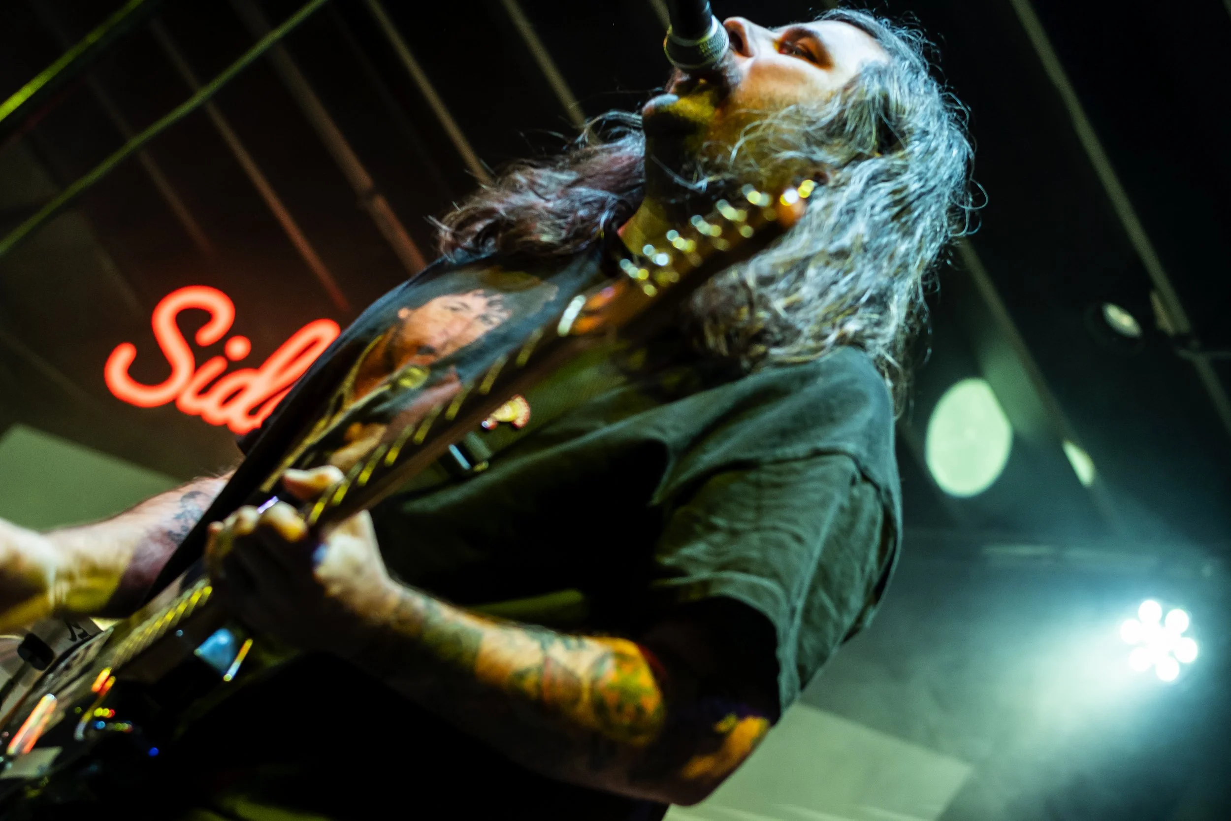 A musician with long, curly hair and tattoos is performing on stage, singing into a microphone, with a neon red sign reading 'Sid' in the background.