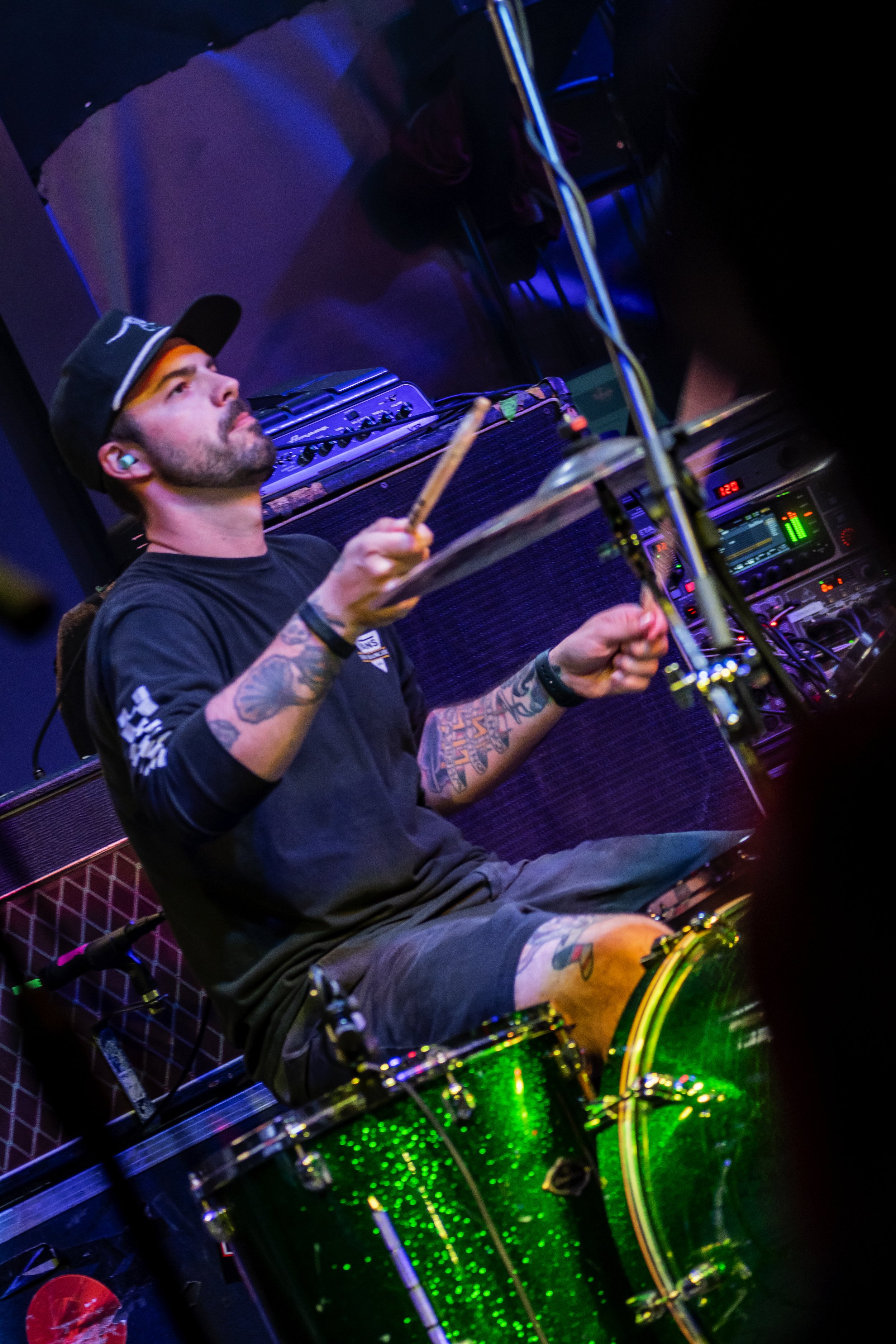 A man with tattoos and a black cap is playing drums in a dimly lit setting with blue and green lighting.