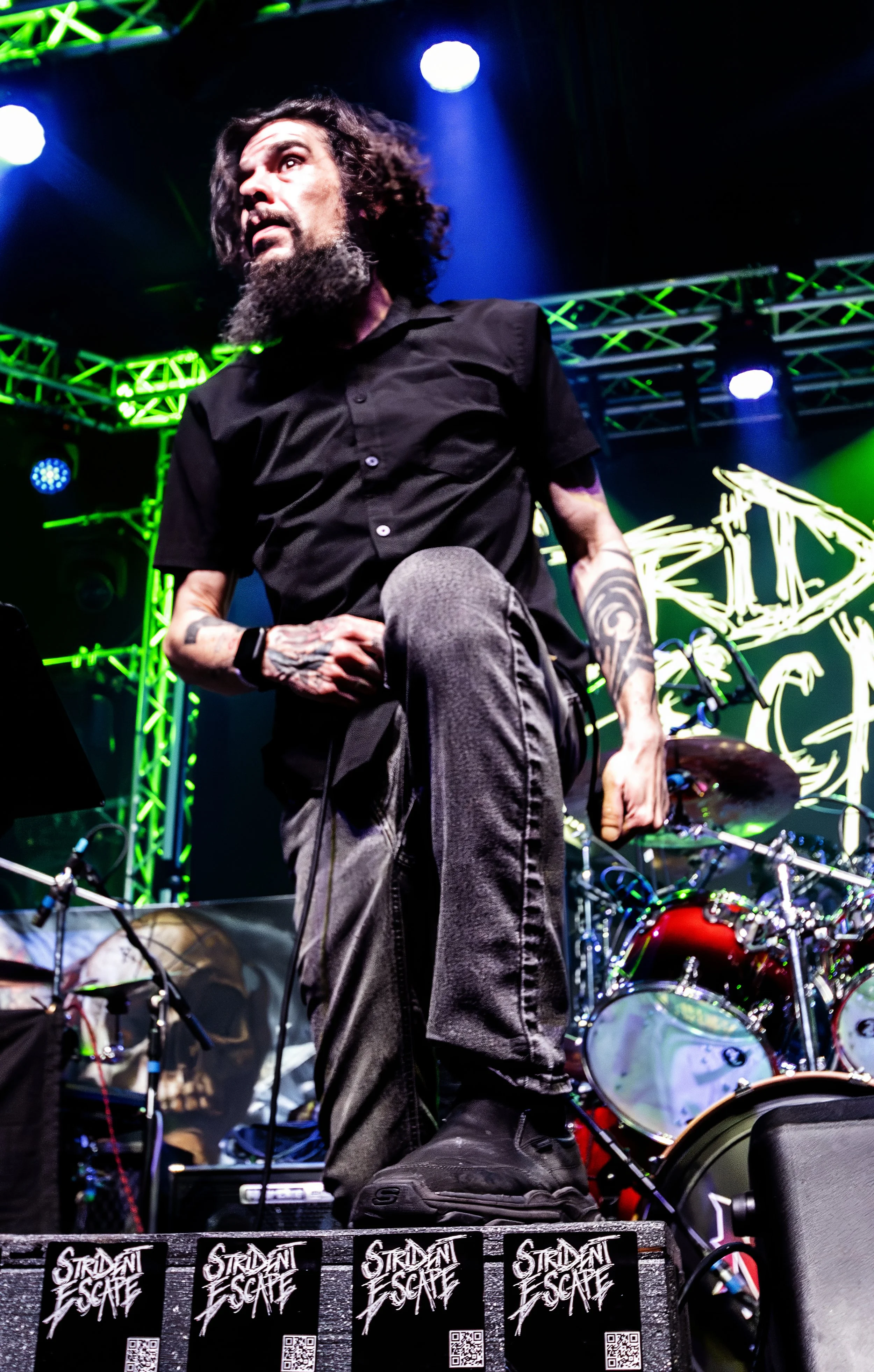A male musician with long curly hair, a beard, and tattoos on his arms performs on stage with drums and green stage lighting in the background.
