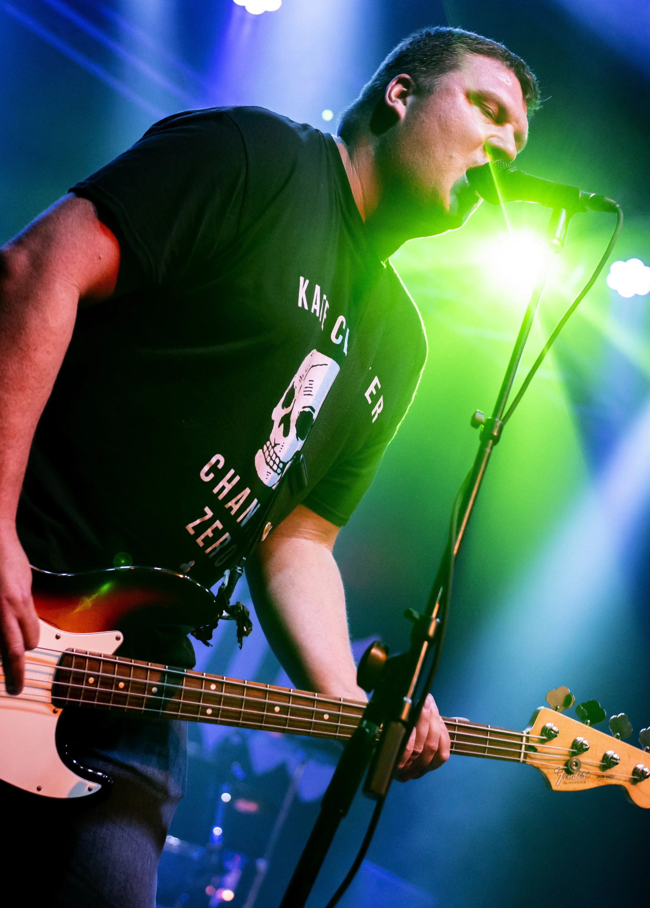 A male musician singing into a microphone while playing an electric bass guitar on stage, illuminated by colorful stage lights.