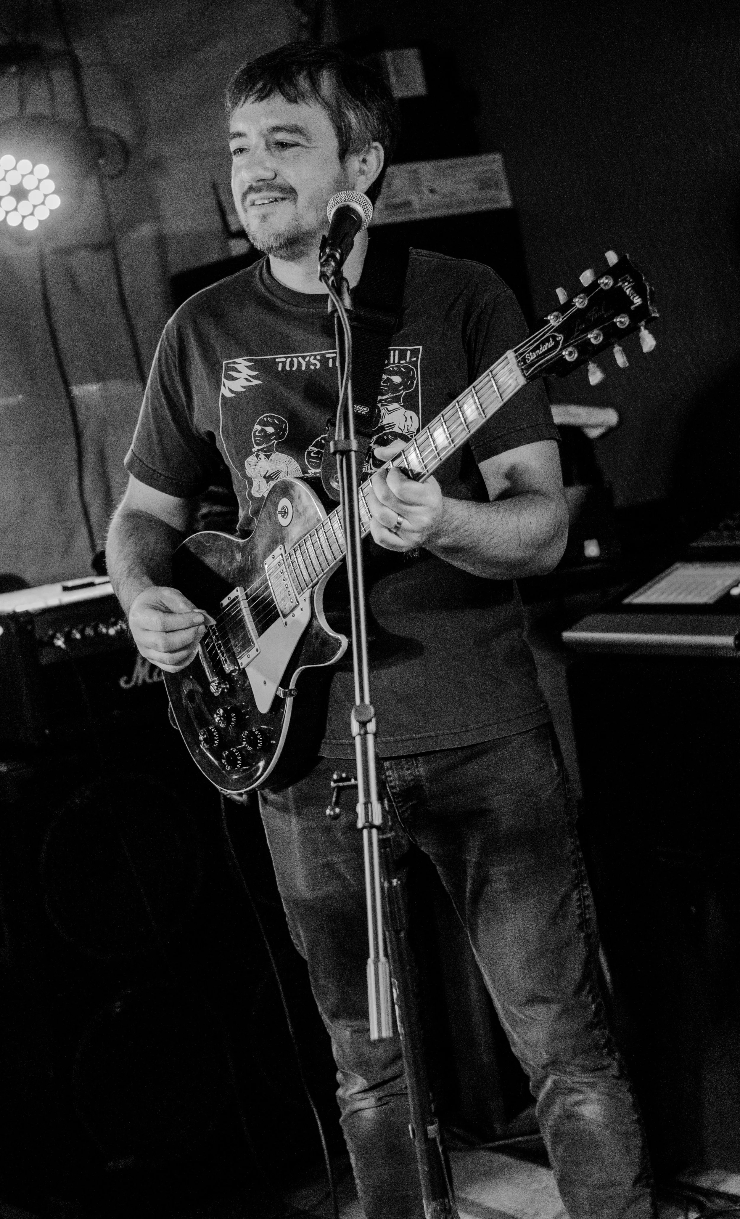 A man with dark hair and a beard playing an electric guitar and singing into a microphone on a stage, in a dimly lit venue.