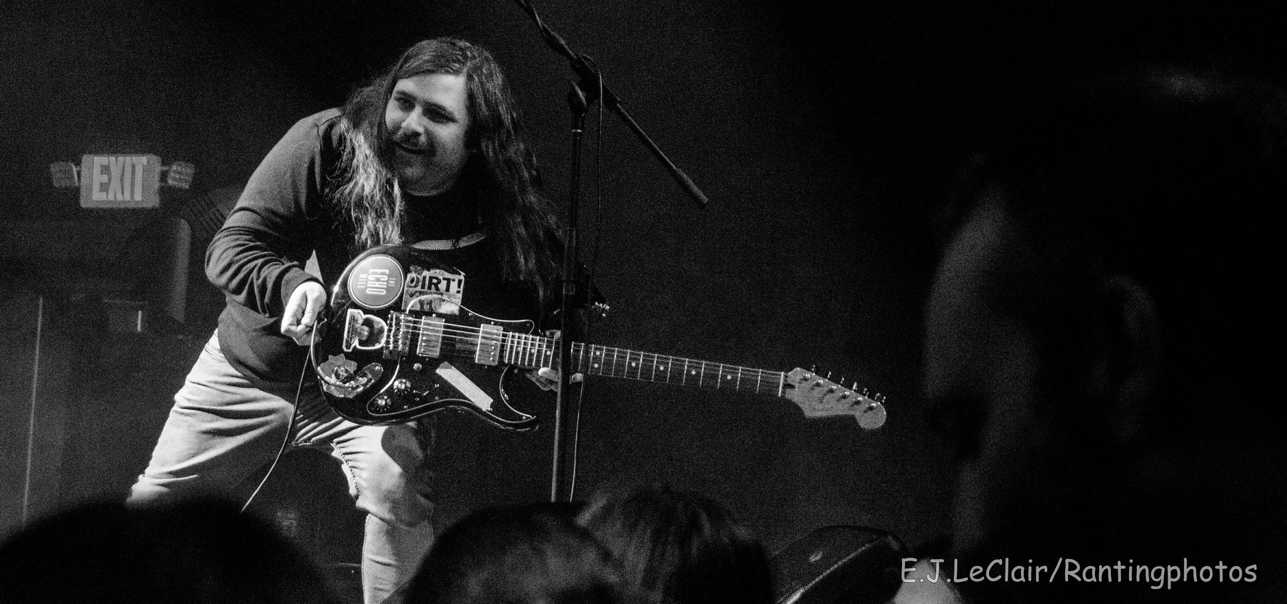 A person with long hair playing an electric guitar on stage, facing slightly to the left, with an 