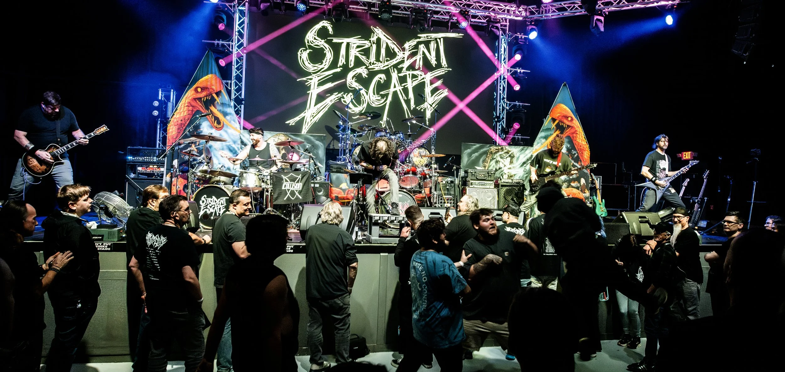 A live music concert with a band performing on stage, titled 'Strident Escape', with people dancing and enjoying the show.