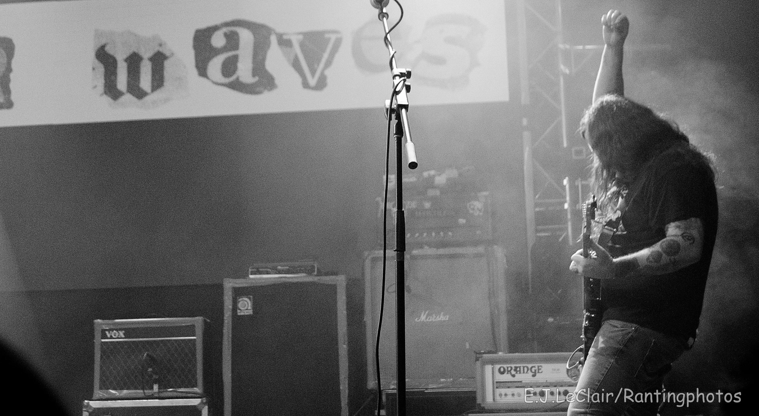 A musician with long hair and tattoos playing an electric guitar on stage, with guitar amplifiers and equipment behind them, in black and white.