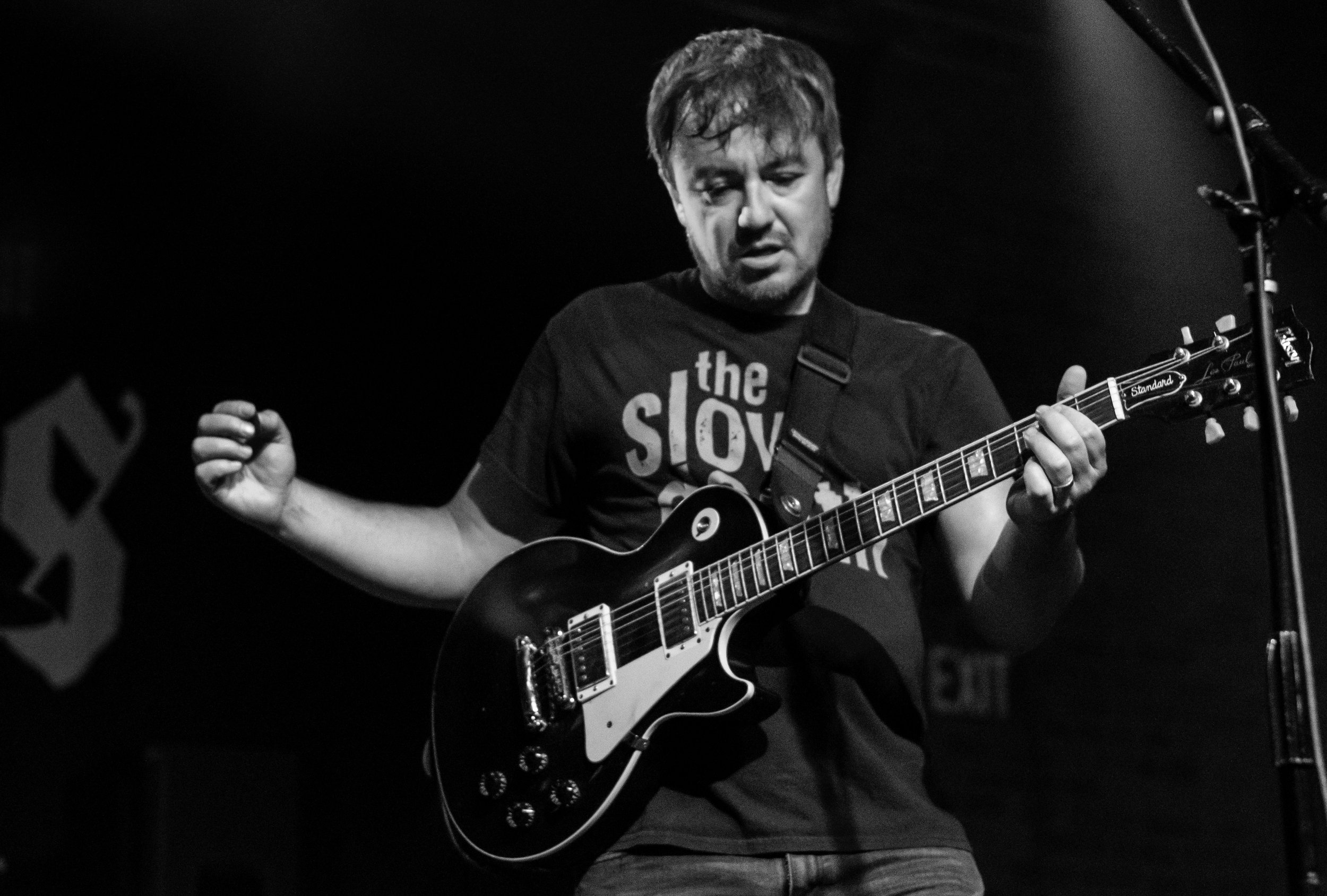 A man playing an electric guitar on stage, with closed eyes and a focused expression, under a dark background.
