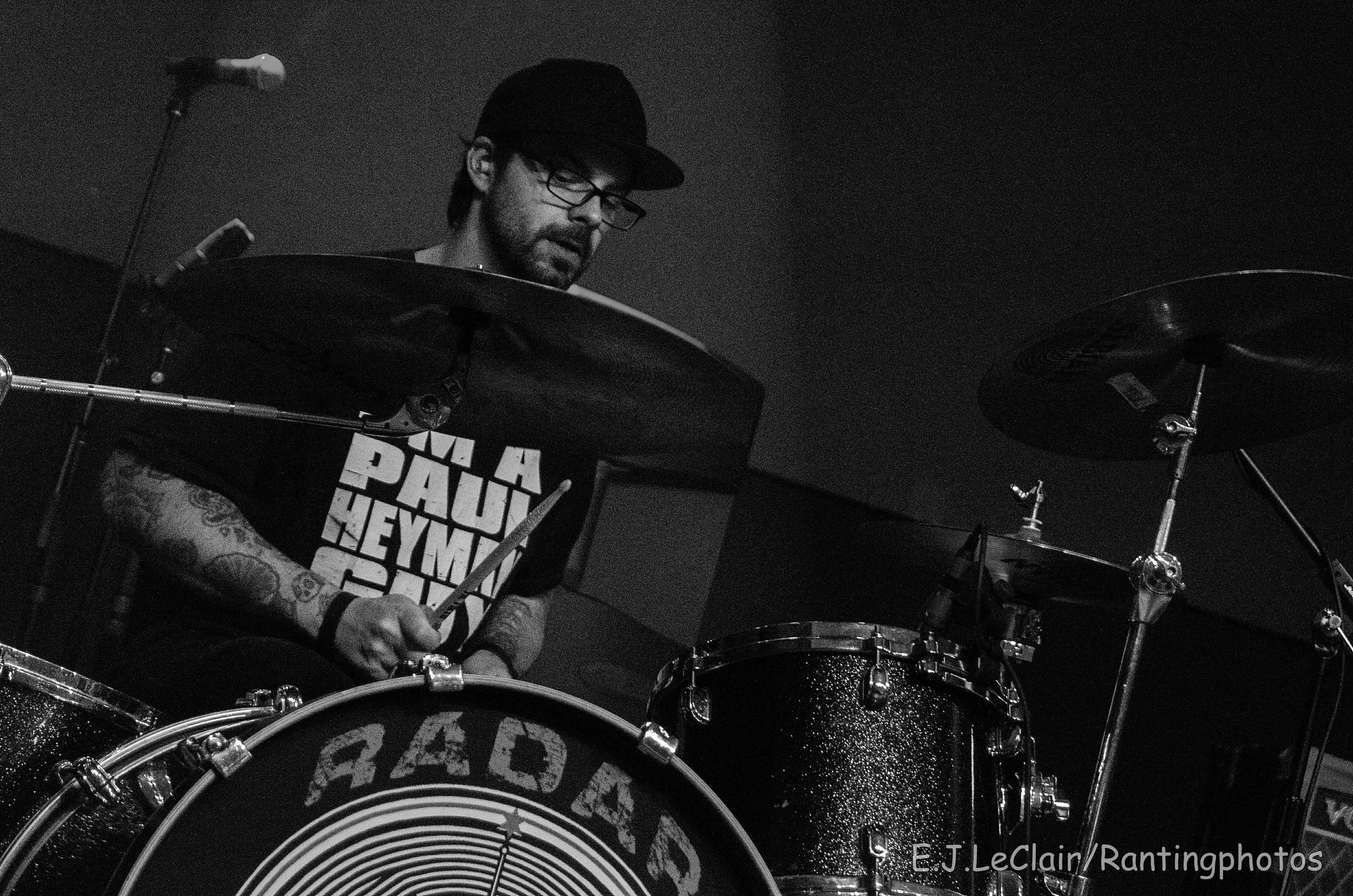 A man with glasses and a cap playing a drum set in a dark room.