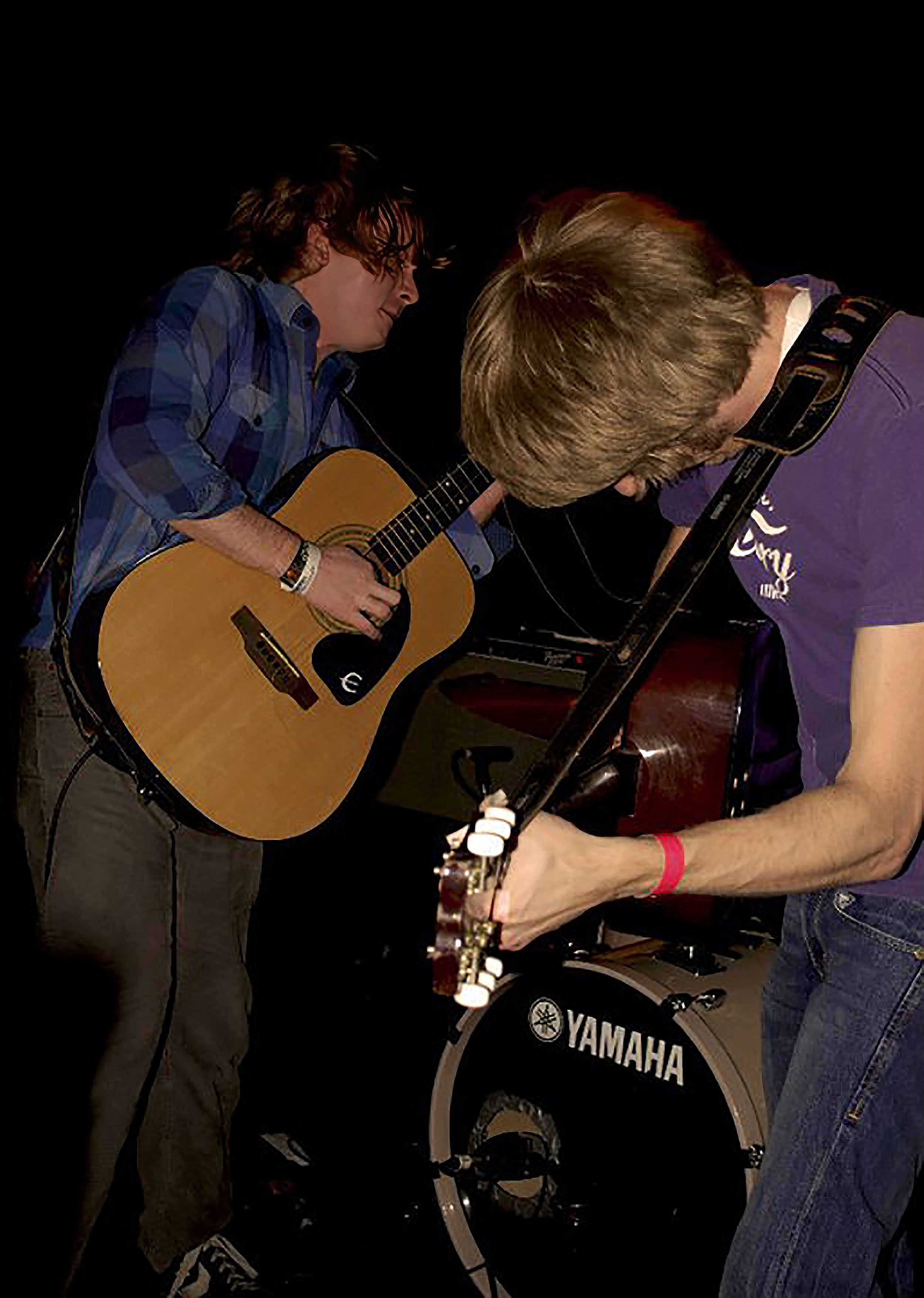 Two young men playing guitars together in a dimly lit setting. One is standing with a yellow acoustic guitar, wearing a blue plaid shirt and dark pants. The other is bent over with a purple shirt, playing a bass guitar. A Yamaha drum set is visible in the background.