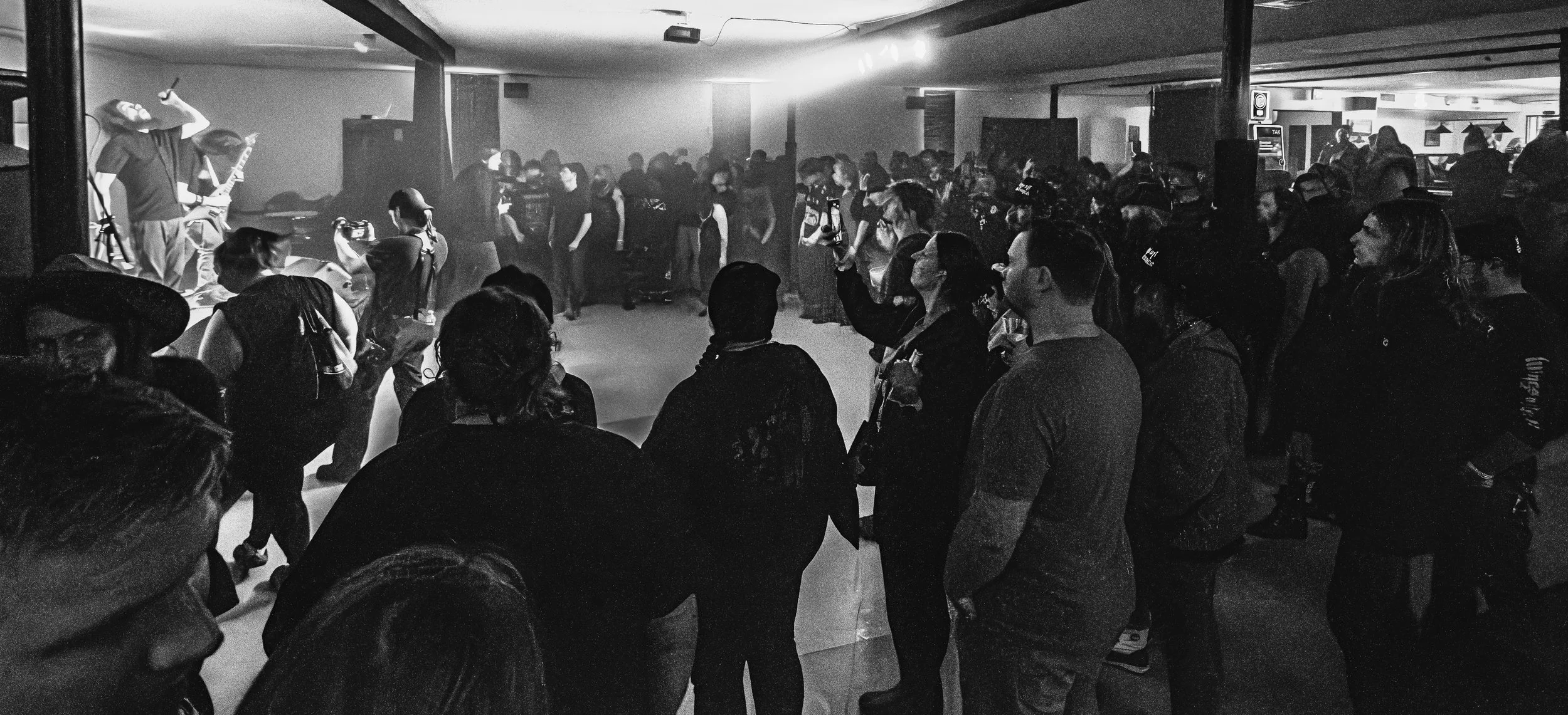 Black and white photo of a music performance in a crowded indoor venue. A guitarist is on stage, with an audience watching intently.