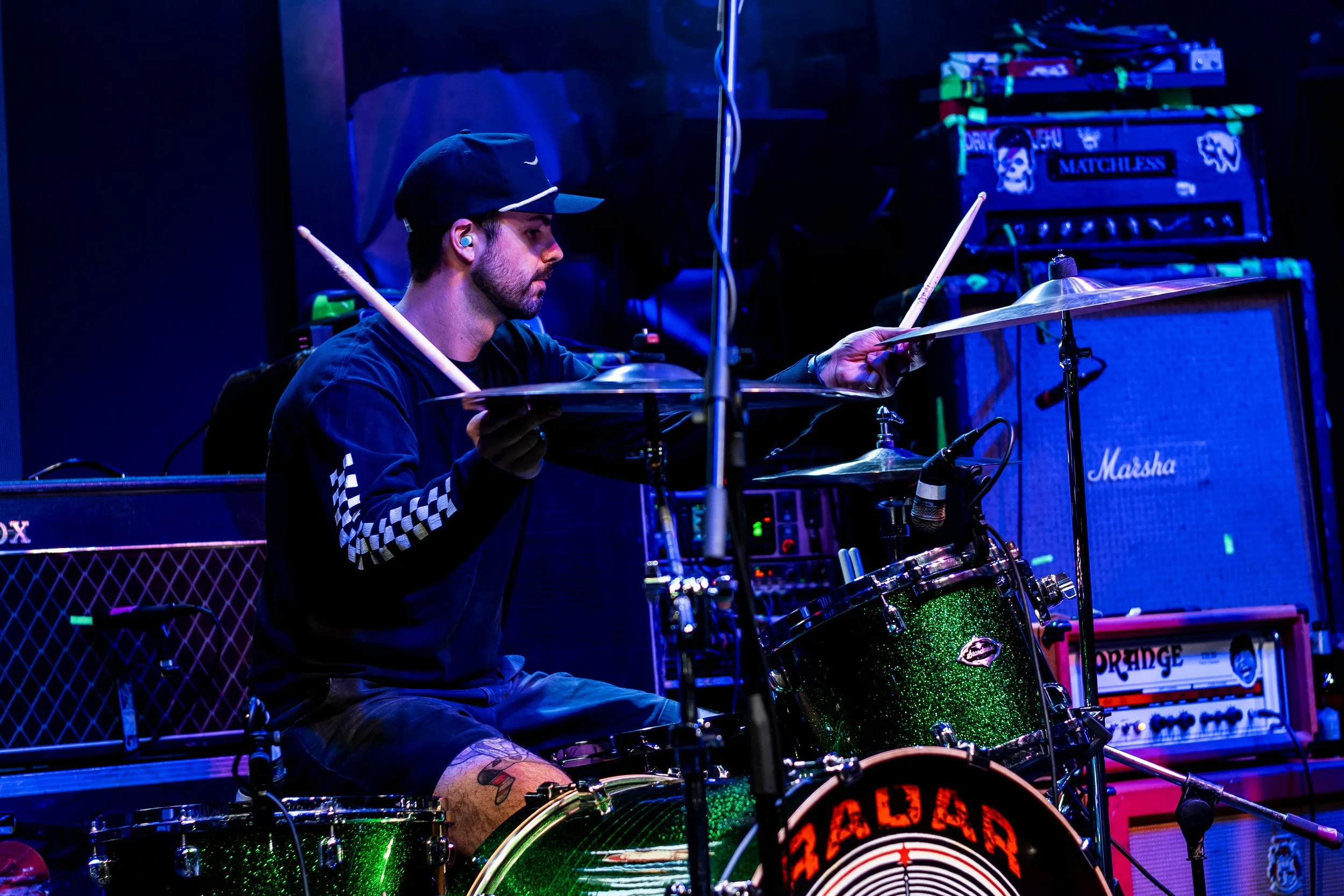 A man wearing a black cap and black jacket with checkered sleeves is playing a drum set on stage, surrounded by music equipment and amplifiers with colorful lights.