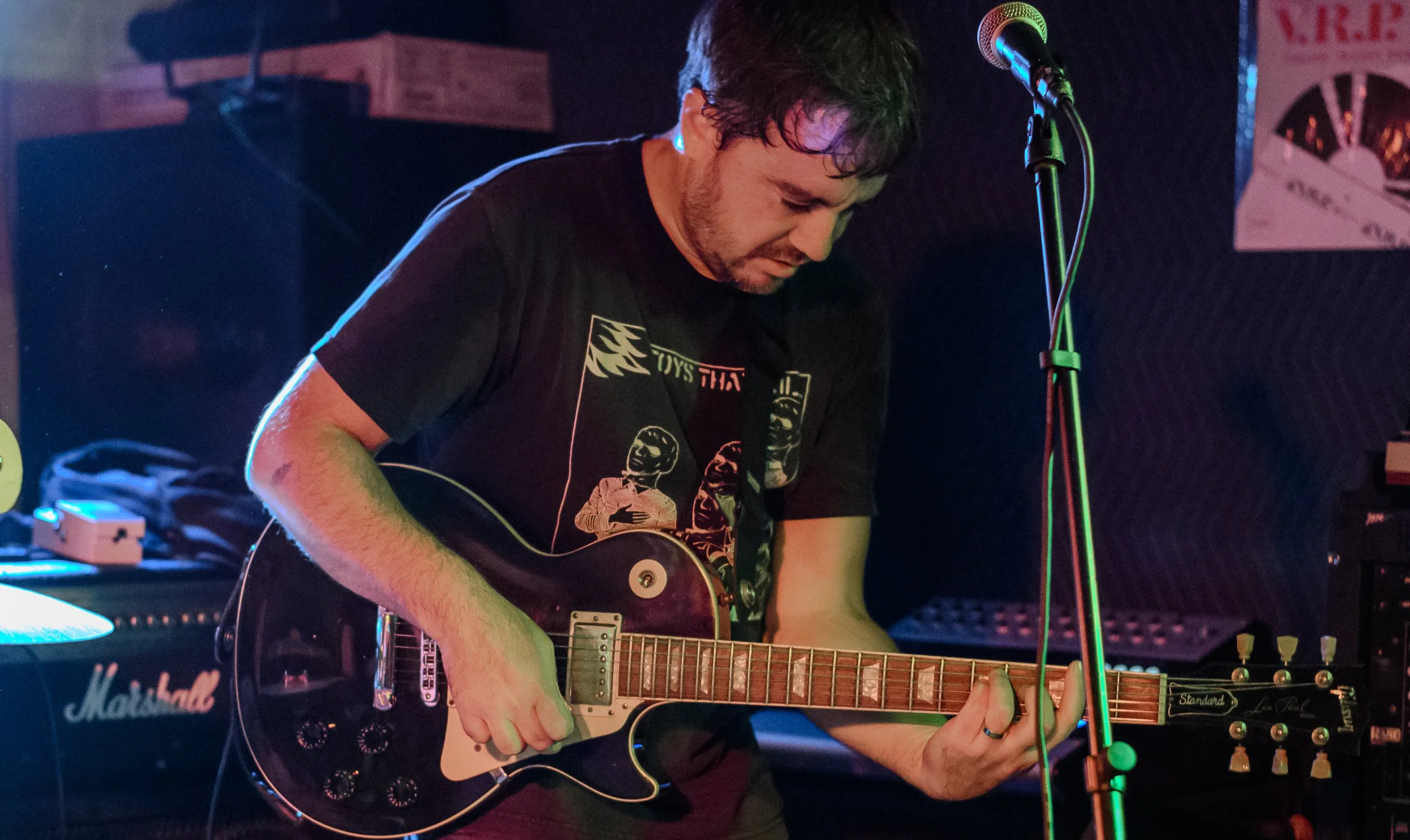 A male musician playing an electric guitar on stage with a microphone nearby, surrounded by music equipment in a dimly lit venue.