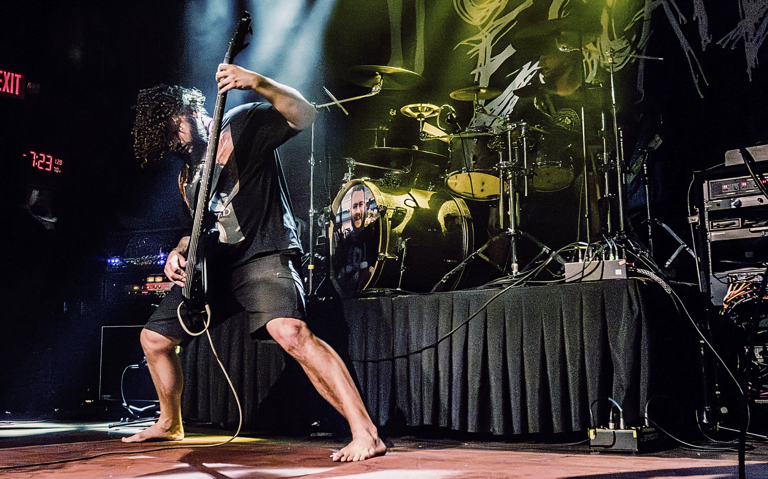 A musician performing on stage, playing an electric guitar with long curly hair, wearing a black T-shirt and shorts, with bare feet. The stage has a drum set and various electronic equipment, and a digital clock shows 7:23. The background is dark wit