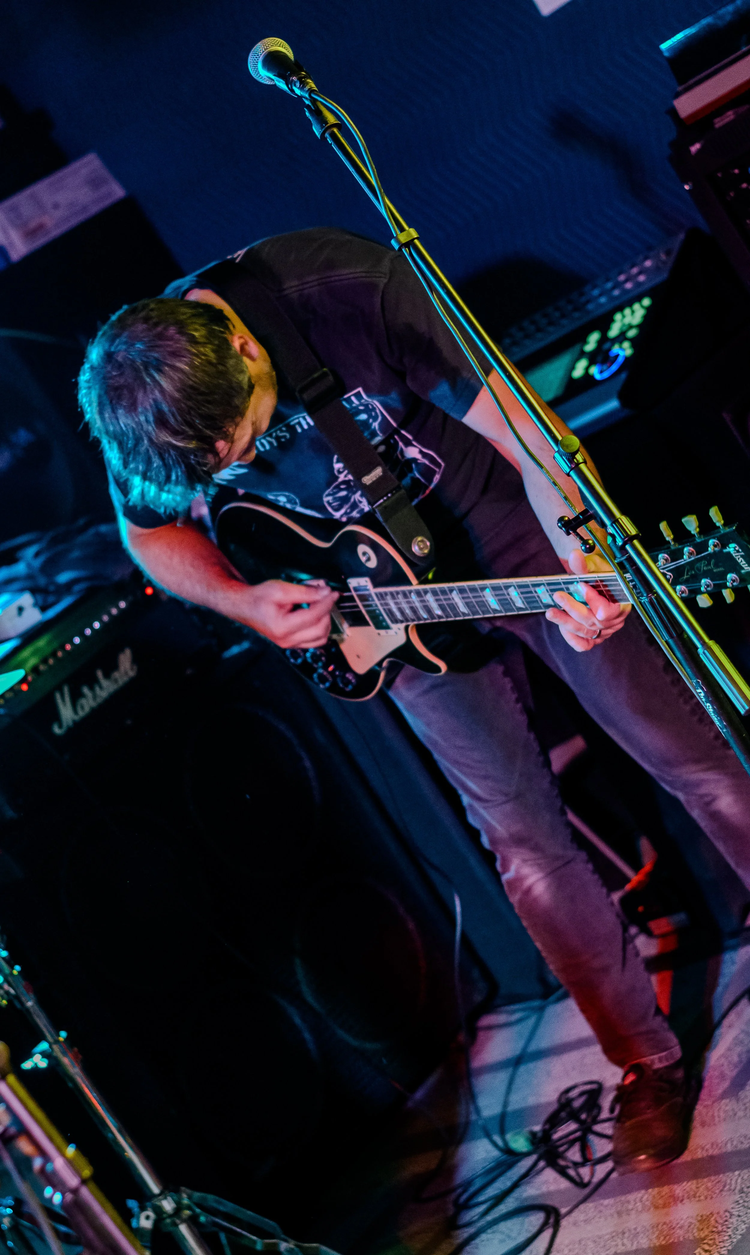 A musician playing an electric guitar on stage with a microphone and amplifier in the background, with stage lighting creating colorful effects.