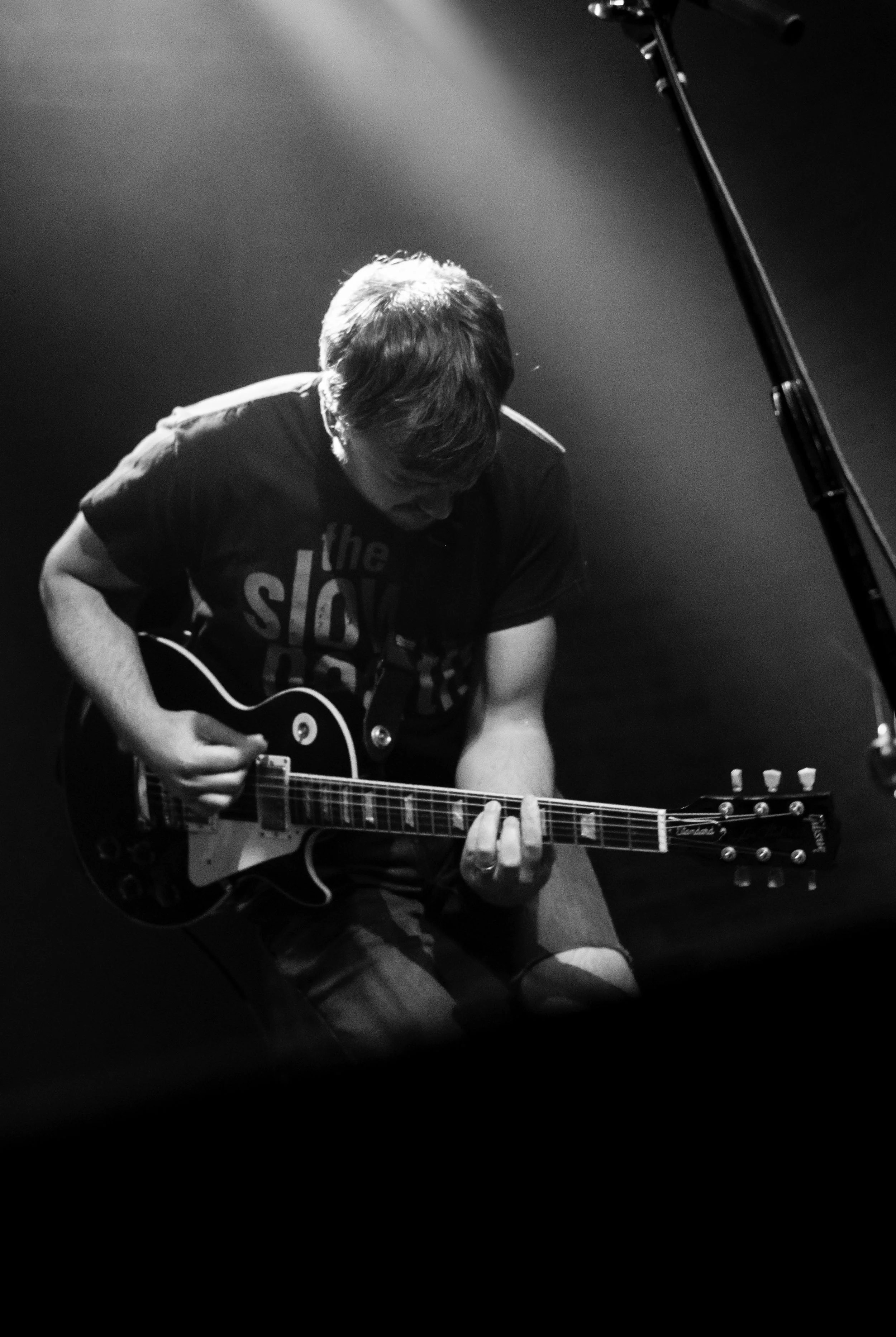 A person sitting on the floor playing an electric guitar, with their head bowed and hair covering part of their face, illuminated by a focused light in a dark setting.