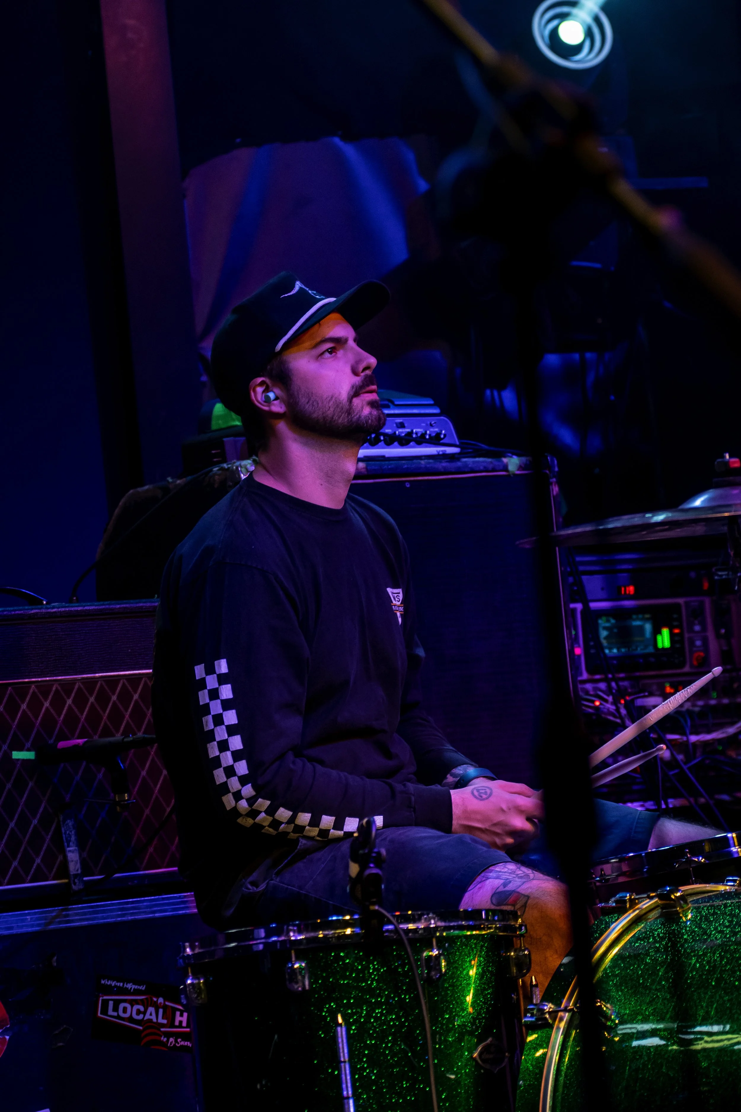 A man with a beard, wearing a black long-sleeve shirt with checkered stripes on the sleeves, shorts, and a black cap, sitting behind a green glittery drum set. He is holding drumsticks and looking attentively upward. The background shows musical equi