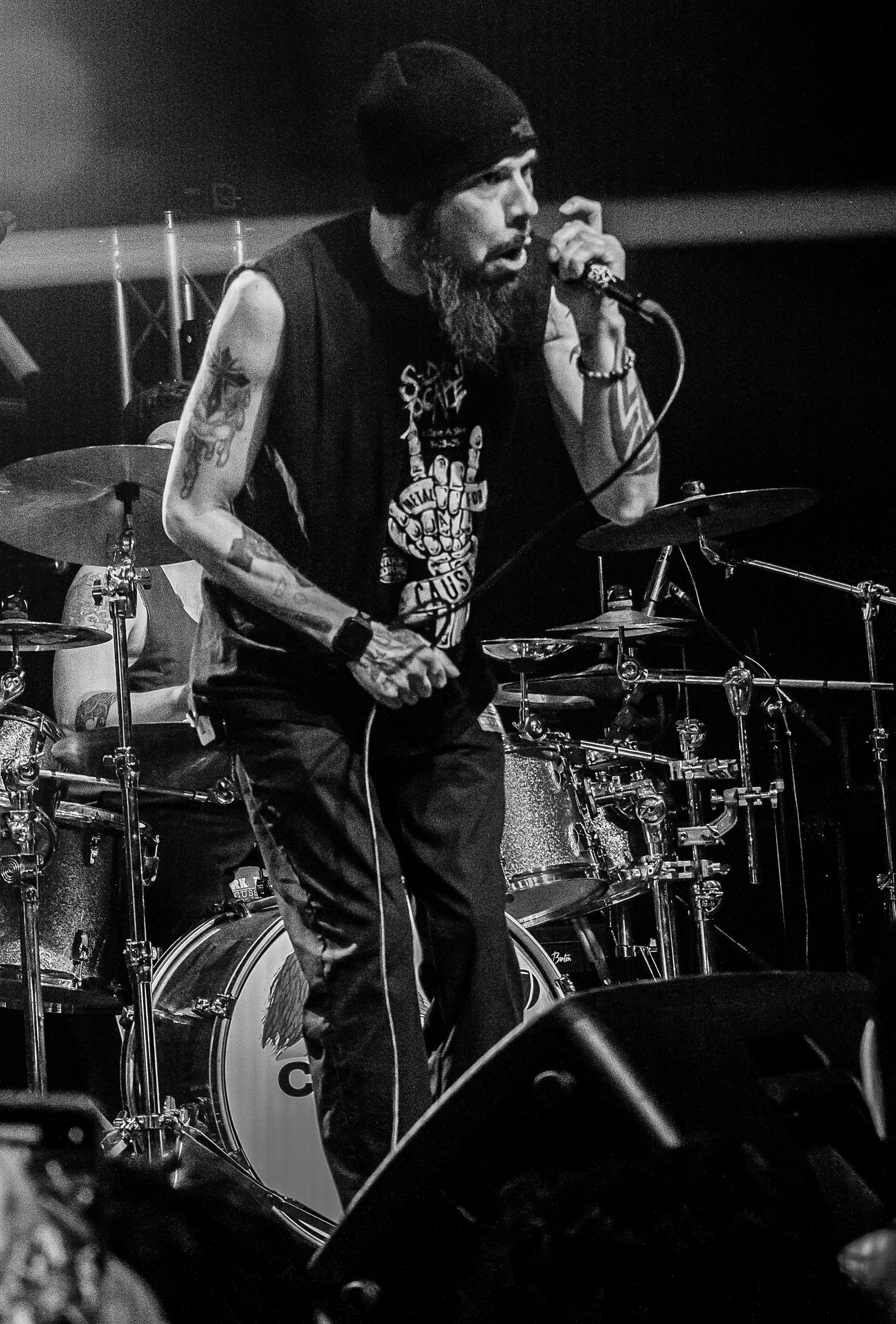 A male drummer with a beard, tattoos, wearing a sleeveless shirt, playing drums and singing into a microphone during a live music performance in black and white.