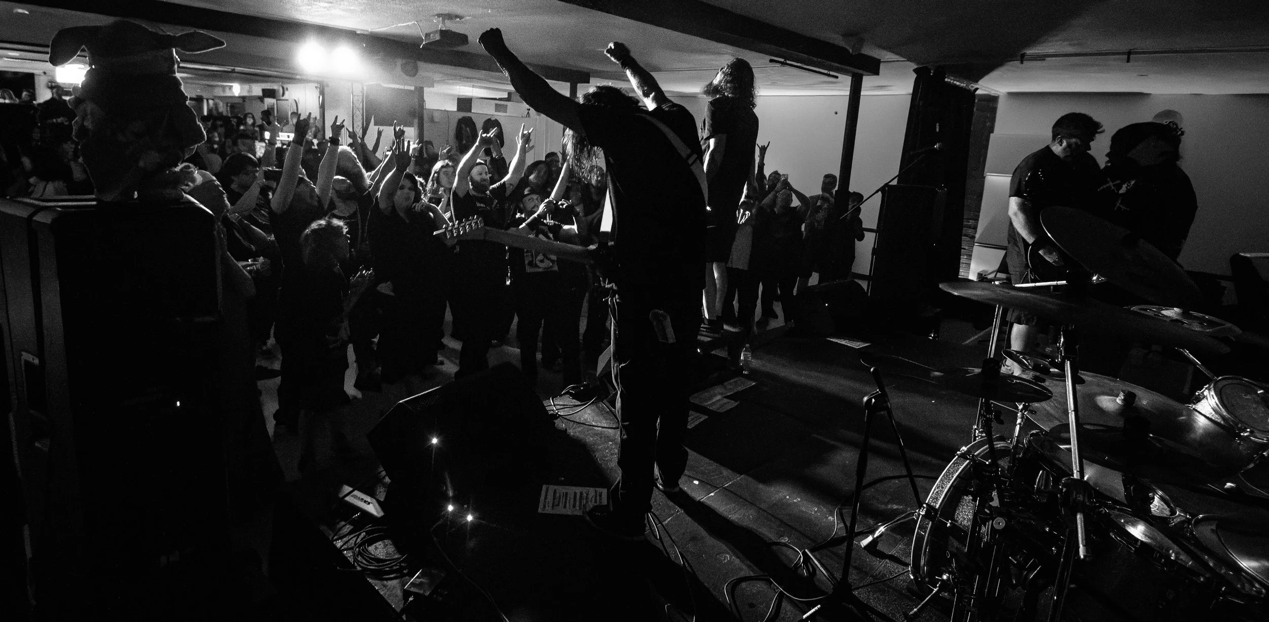 Black and white photo of a live music concert in a small venue. The audience is raising their hands, and band members are playing on stage with one person holding a guitar. Stage lights are illuminating the scene, creating a lively atmosphere.