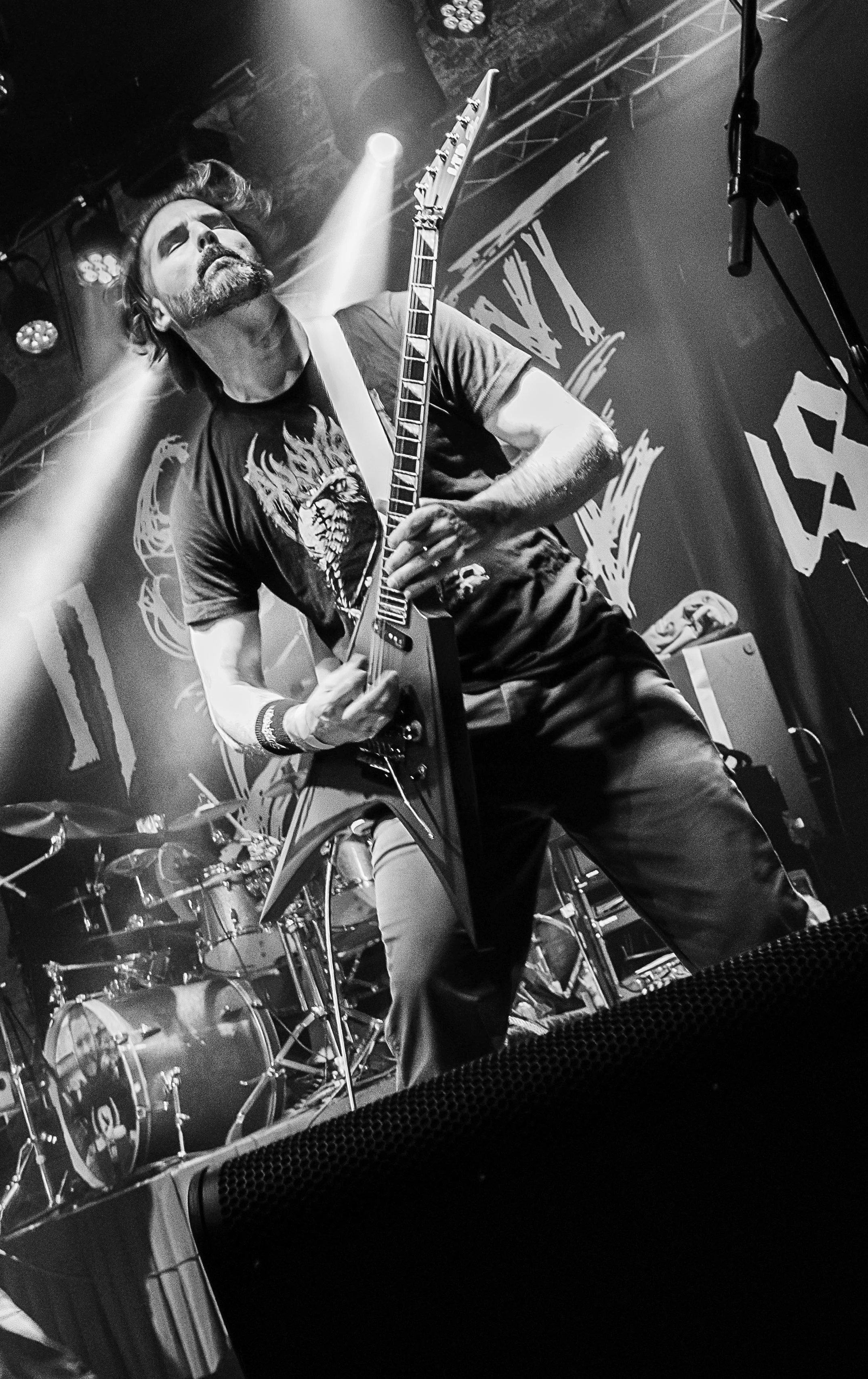 Black and white photo of a male guitarist with a beard and styled hair, performing on stage with drums in the background and stage lights shining down.