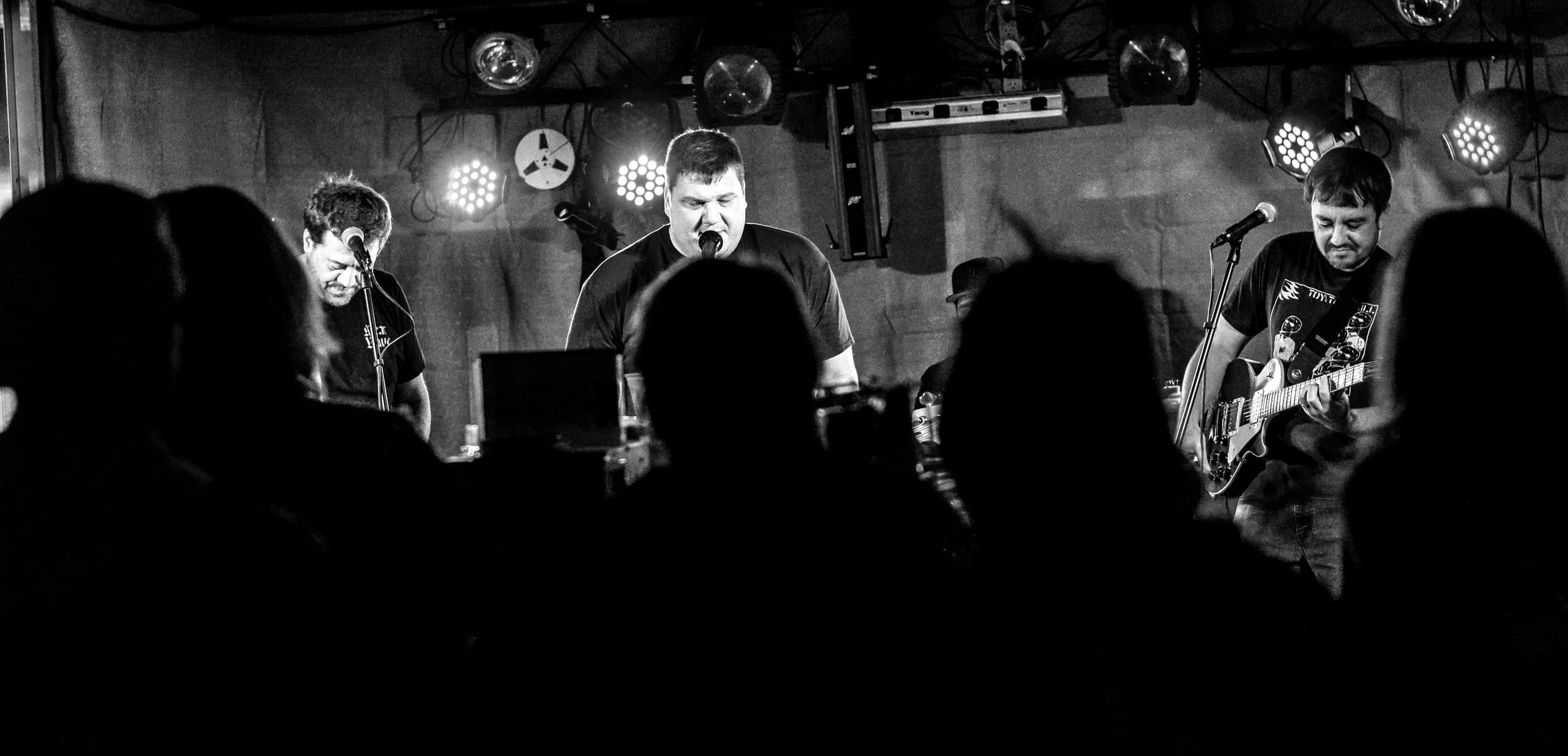 Black and white photo of a band performing on stage with three members, one singing and playing guitar, two with microphones, in a dimly lit venue with stage lights and audience shadows in the foreground.
