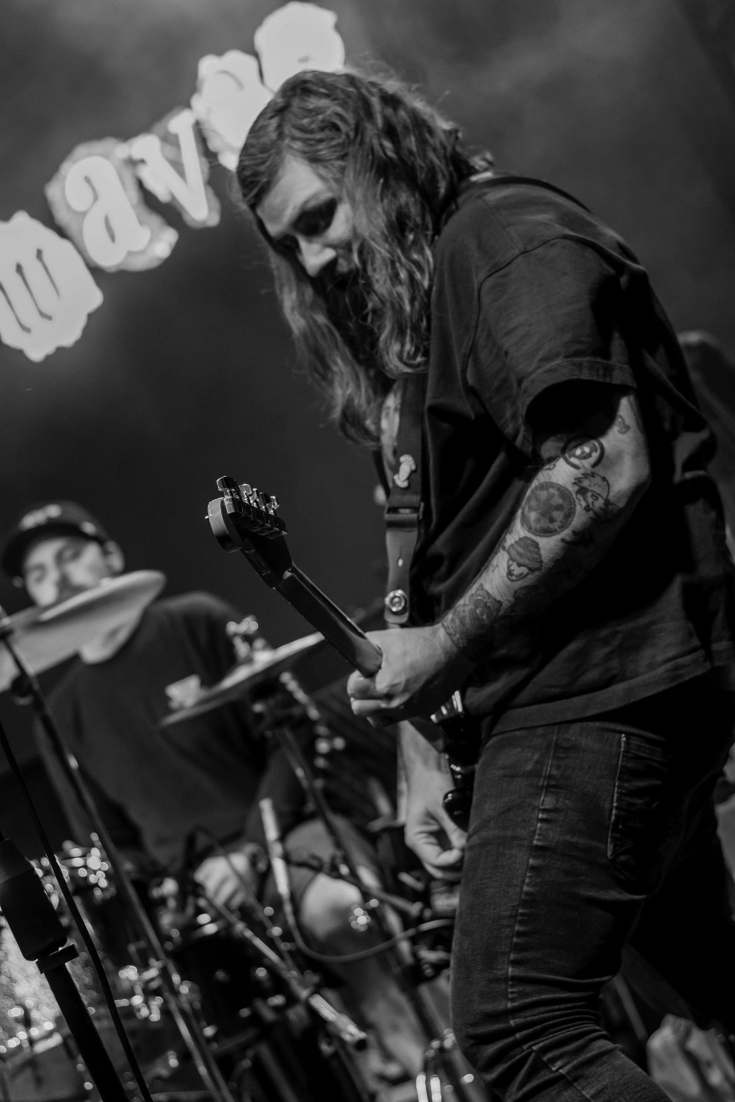 A man with long hair and tattoos playing an electric guitar on stage, with a background of blurred audience and band members.