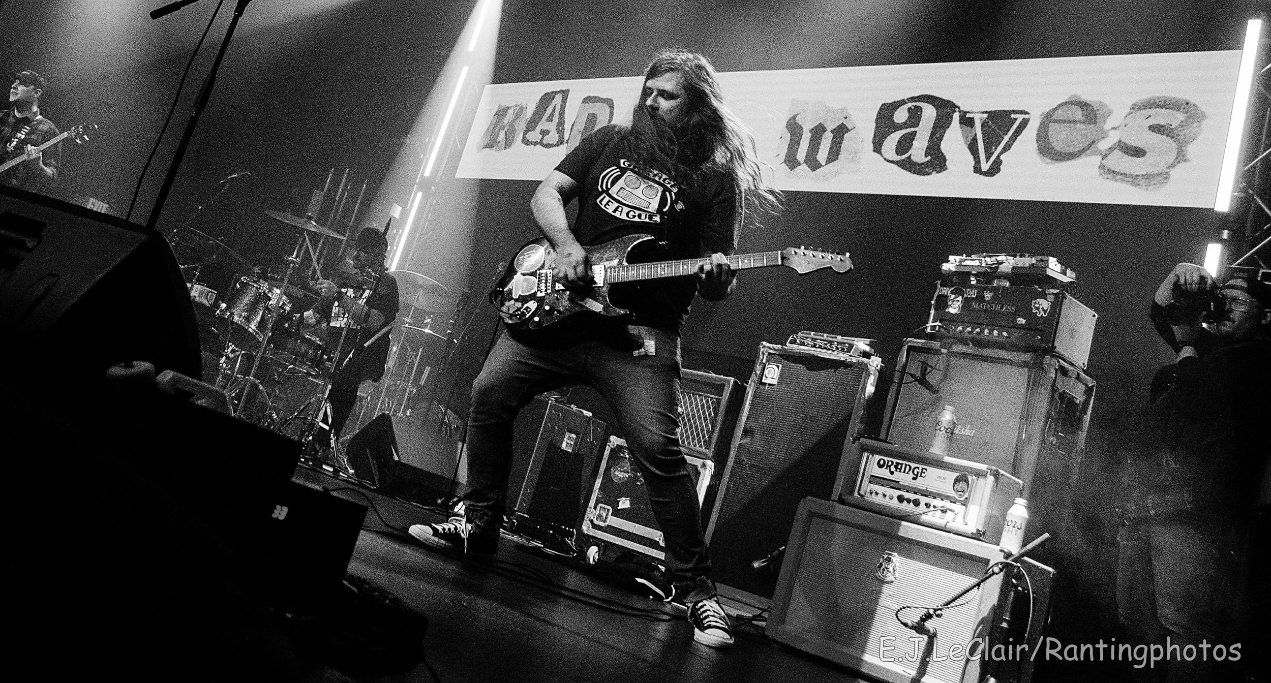 Black and white photograph of a live concert. A long-haired guitarist is playing an electric guitar on stage, surrounded by amplifiers and musical equipment. There are other band members and stage lights, with a large banner in the background that sa