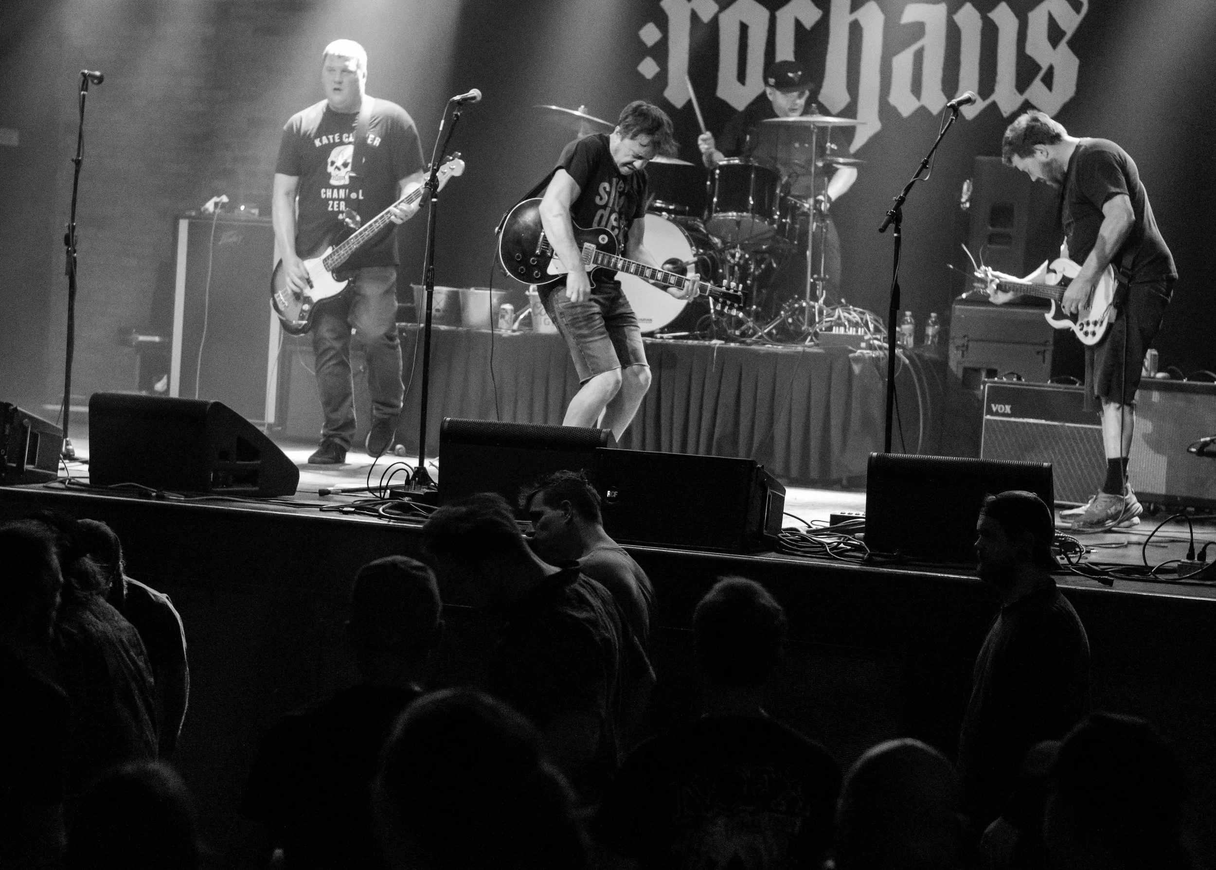 A black and white photo of a live band performing on stage with four members playing instruments, including electric guitars and drums. Audience members are visible in the foreground.