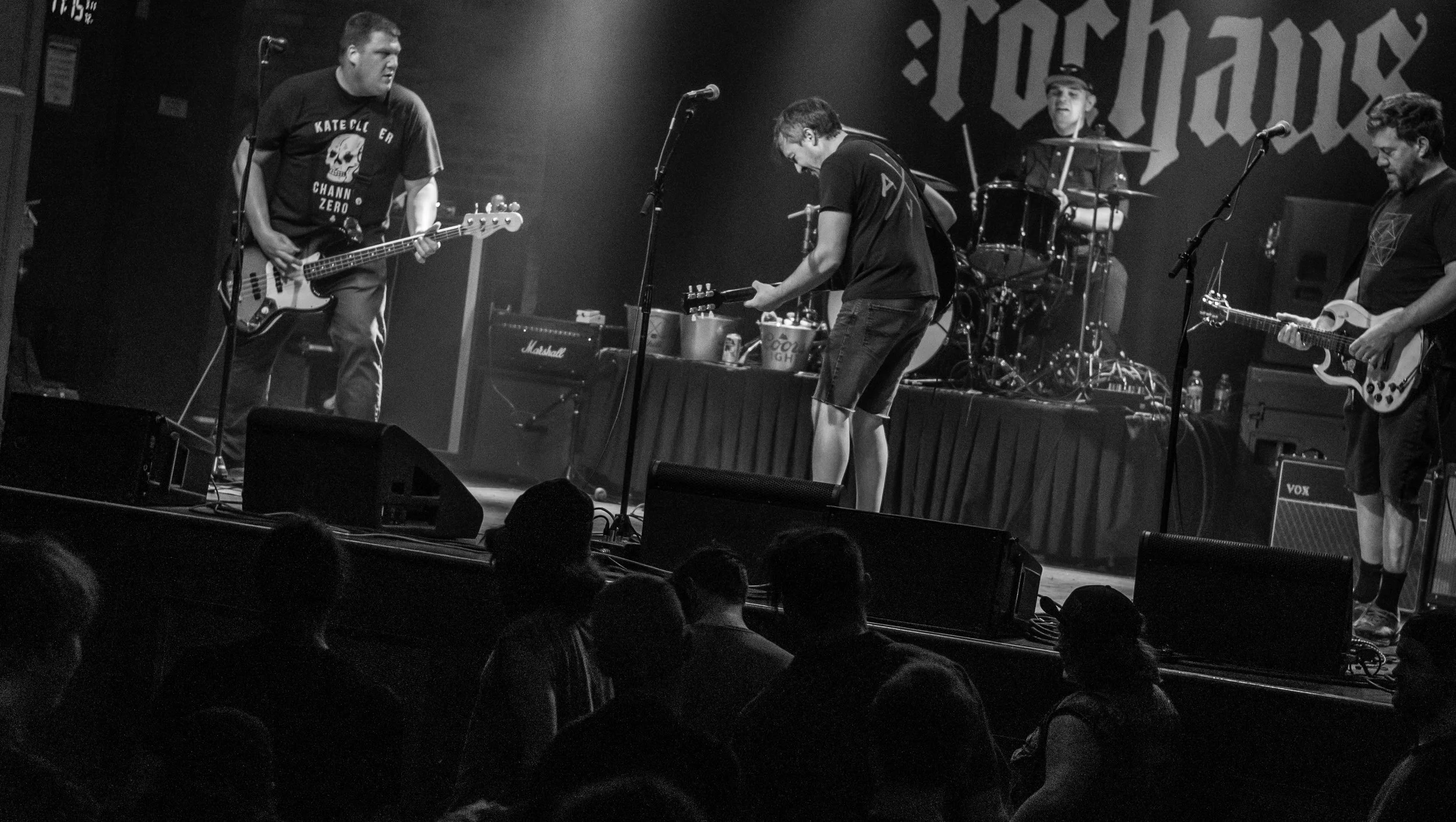 A black and white photo of a band performing on stage with four members playing guitars and drums, with audience members visible in the foreground.