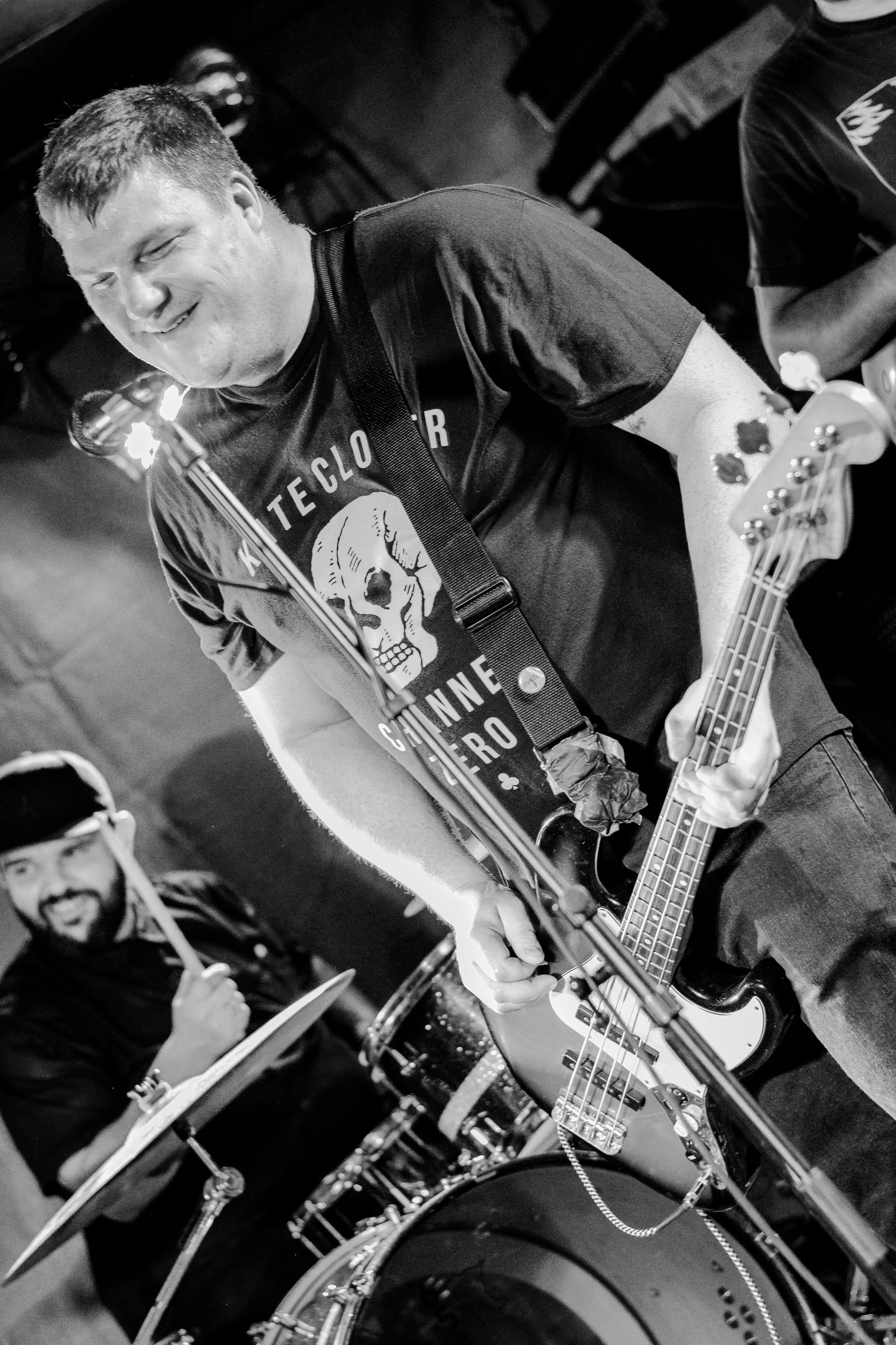 Black and white photo of a smiling musician playing an electric guitar, wearing a t-shirt with a skull graphic, in a dark indoor setting with another person in the background playing drums.