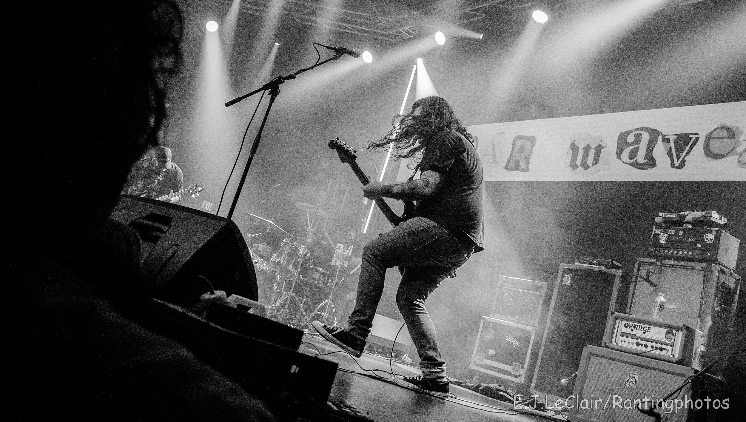 A musician with long hair playing an electric guitar on stage during a concert, with stage lights and equipment visible. The image is in black and white.