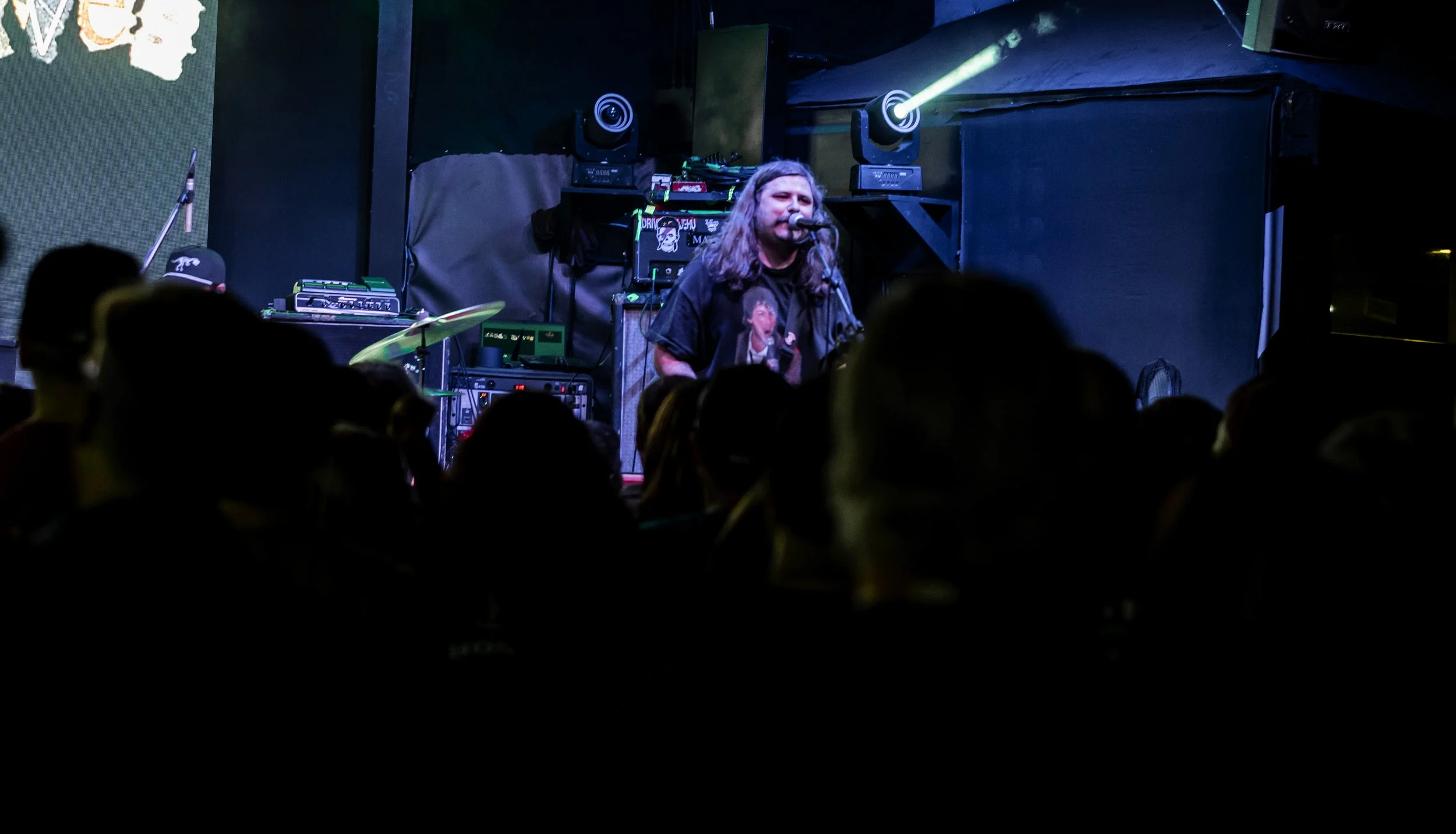 A male singer with long gray hair performing on stage with microphone, dark background, audience silhouette in foreground, stage lighting.