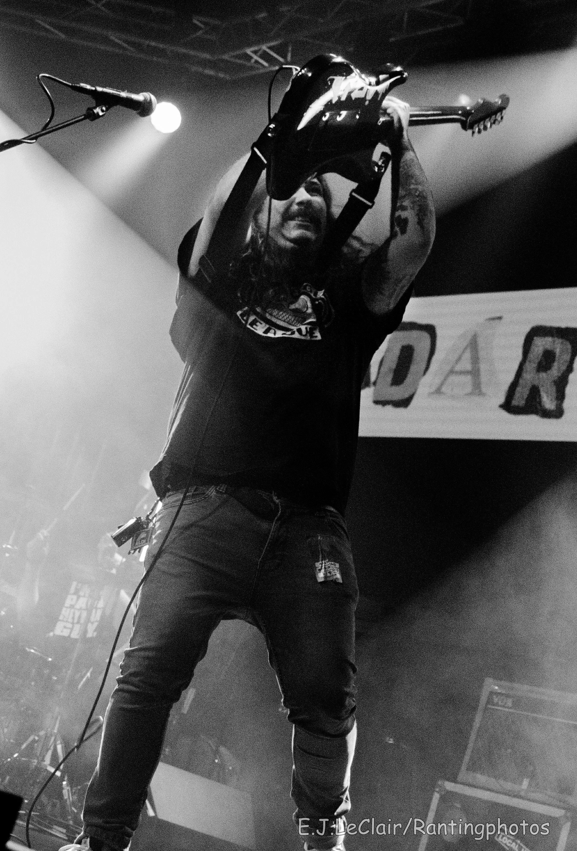 A man on stage holding a guitar above his head during a concert, with a microphone on stage and stage lights. The photo is in black and white.