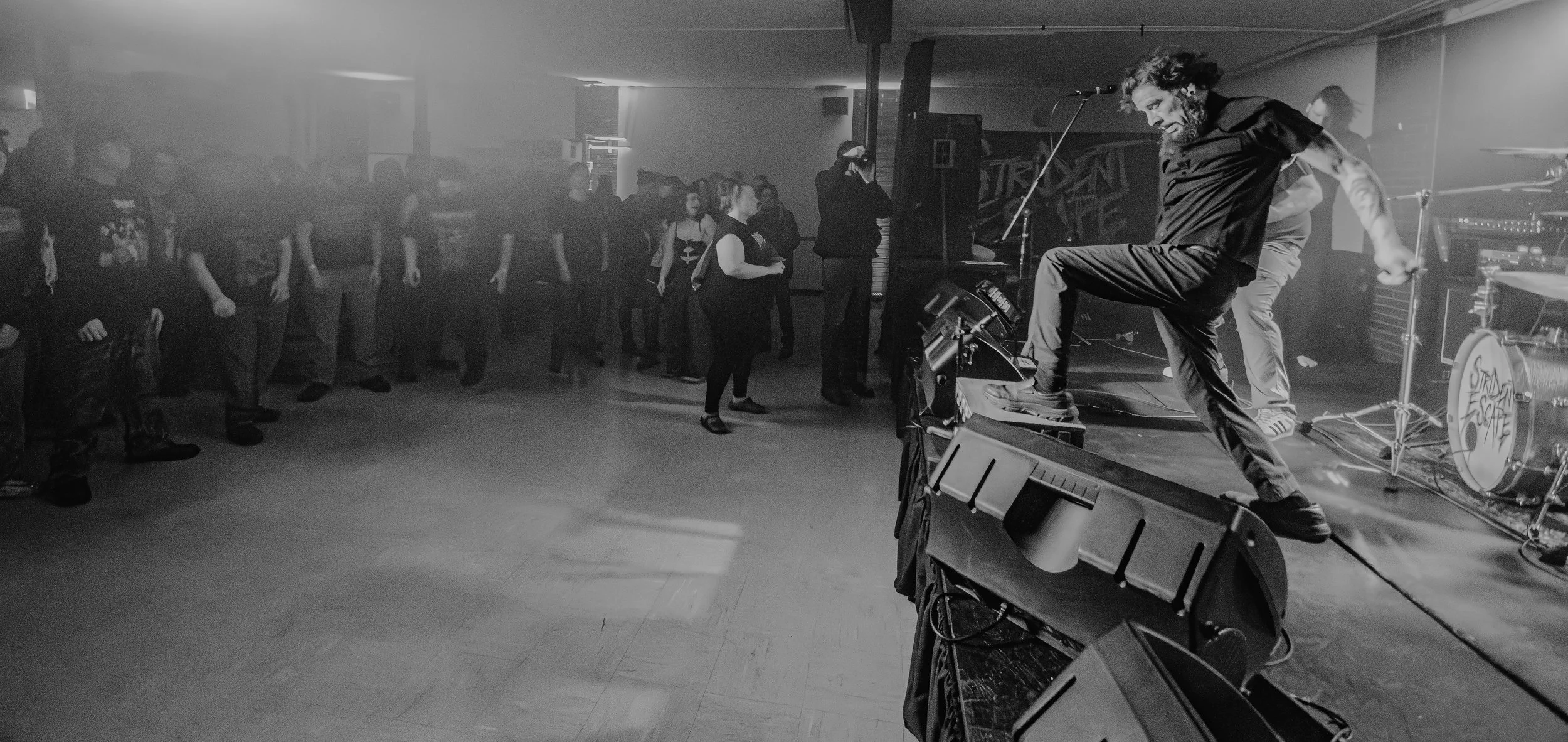 Black and white photo of a live indoor music performance. A man with a beard and long hair is performing passionately on stage with a microphone, raising his leg onto a monitor speaker. Audience members are standing and watching, some with arms cross