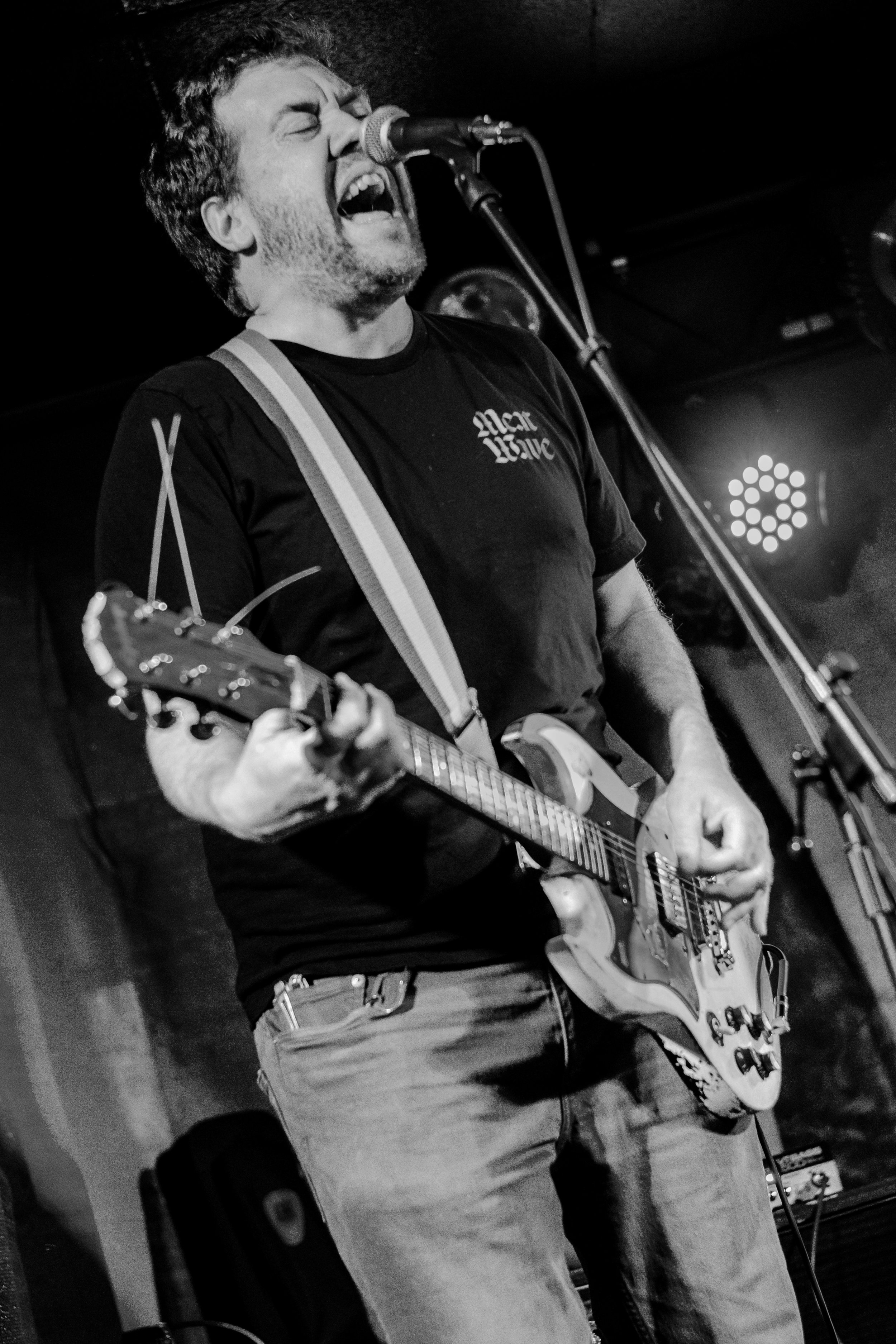 A man with a beard and short hair playing an electric guitar and singing into a microphone on stage. He is wearing a t-shirt and jeans.