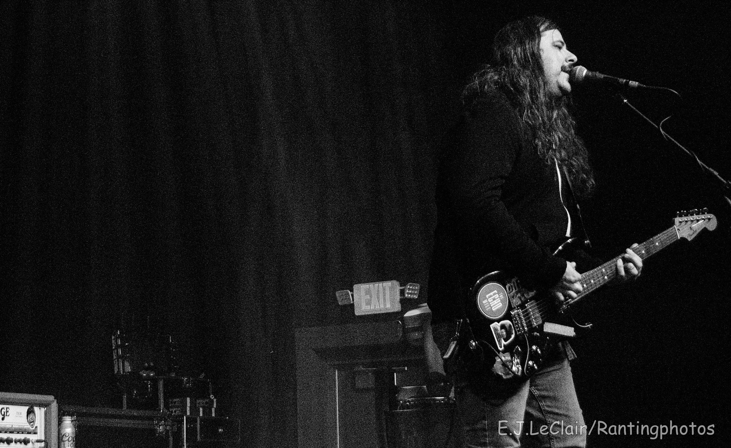 A musician with long hair and a beard singing into a microphone while playing an electric guitar on stage. Black and white photo.