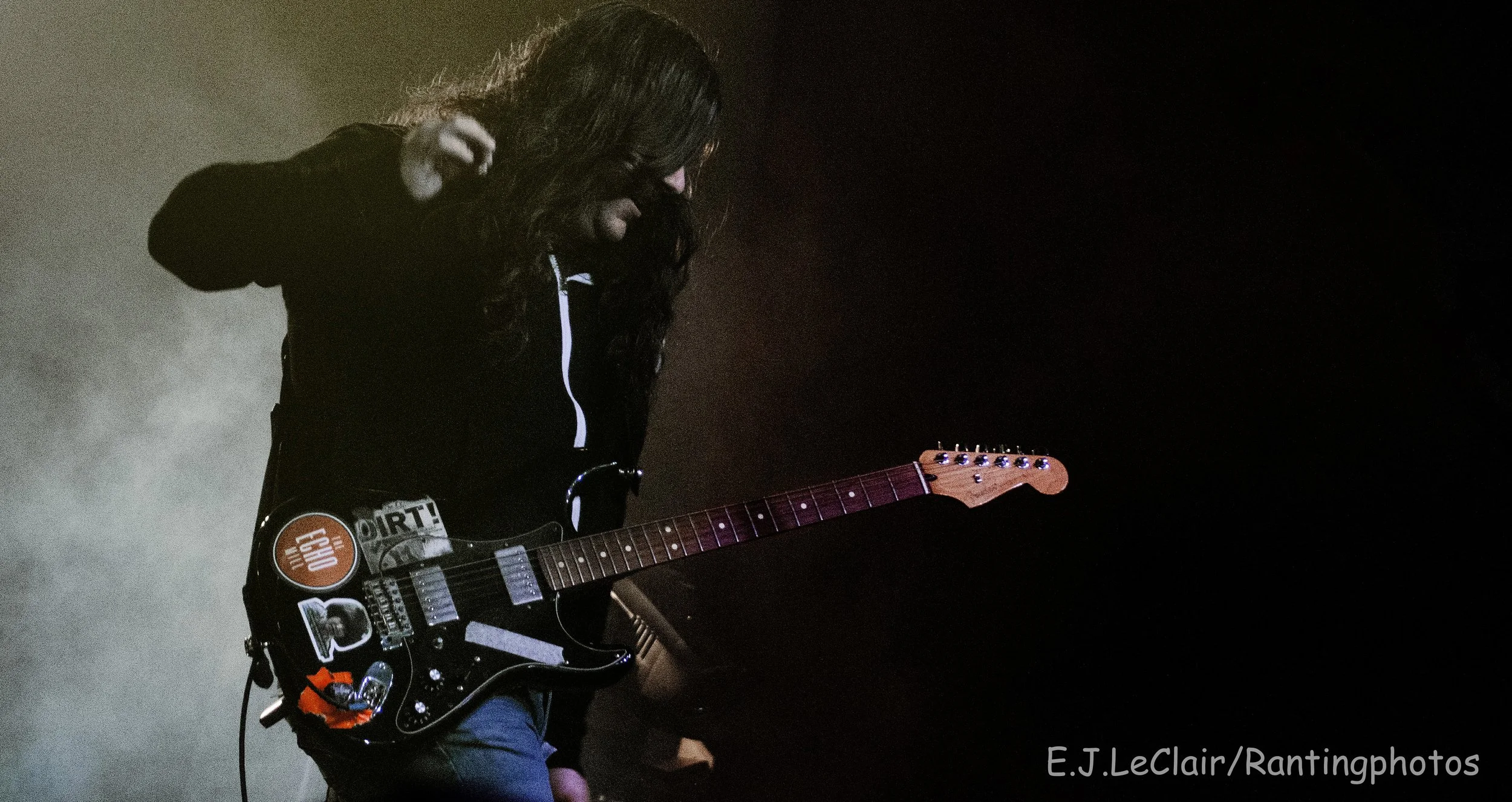 A woman with long dark hair playing an electric guitar on stage, with a dark background.
