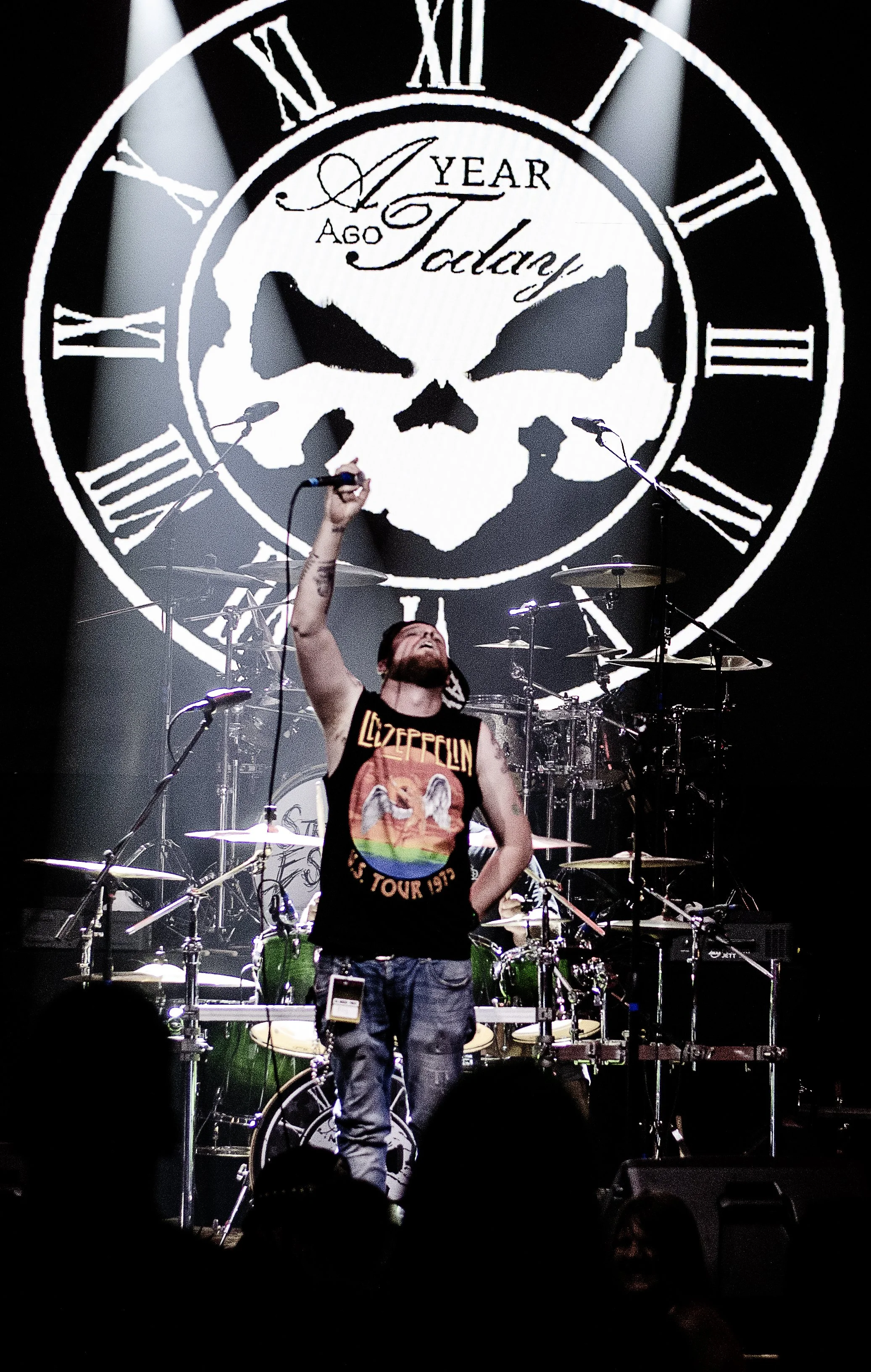 A musician on stage during a concert, wearing a Led Zeppelin t-shirt and jeans, holding a microphone over his head. Behind him is a drum set, and a large screen displays a skull with a clock around it, showing the time on the clock face and text indicating the countdown to a certain year.